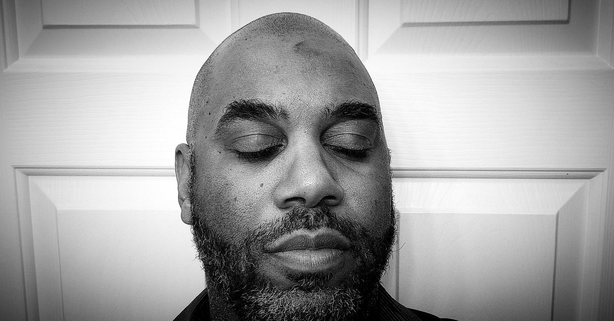 Black and white image of a Black man's face with a shaved head and salt-and-pepper beard. He is standing in front of a white door, his eyes closed, and head turned slightly to one side. A faint smudge of ash is at the center of his forehead.