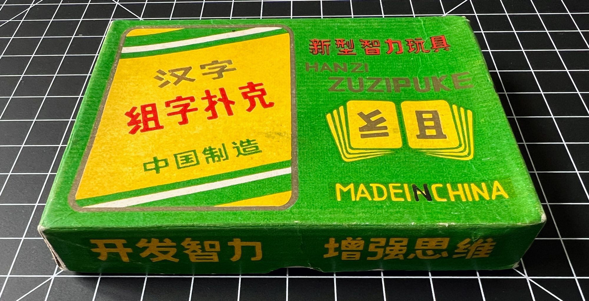 A view of the box package for 汉字组字扑克 (Chinese character combination poker) from the top. The box is green and has various designs combining typography and illustrated playing cards.