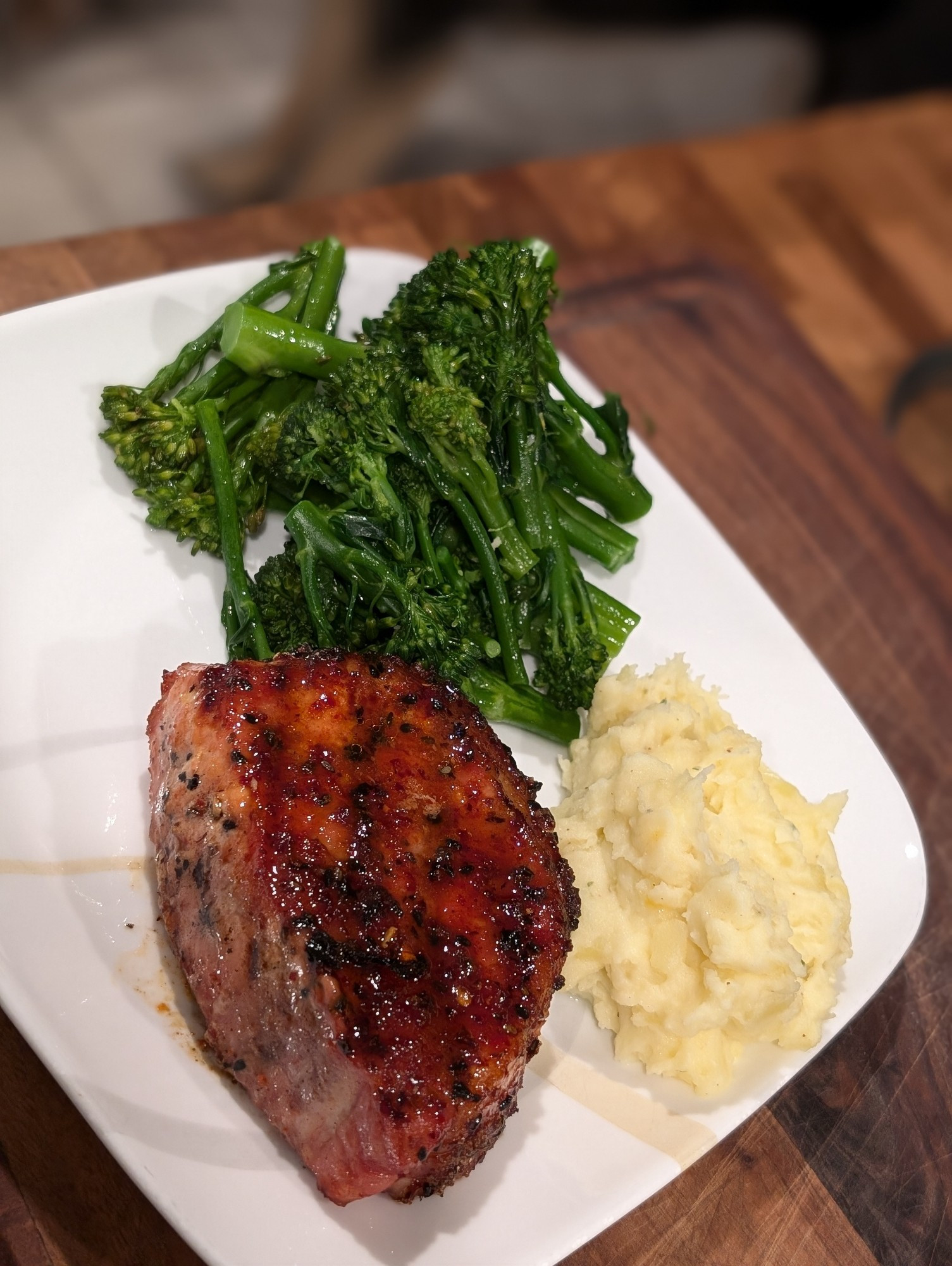 A plate of food with a seasoned and sauced pork chop, broccolini and mashed potatoes 