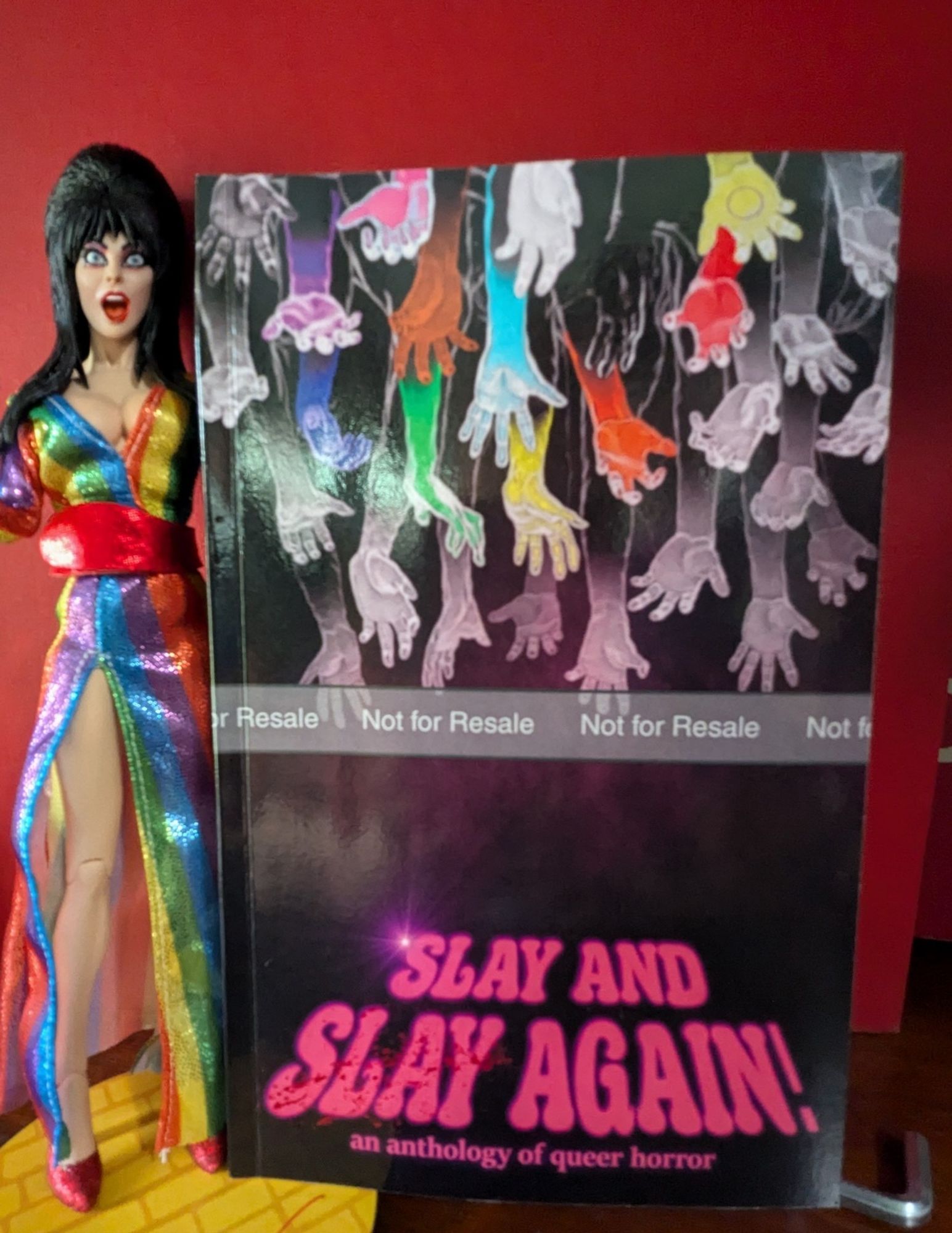 The SLAY AND SLAY AGAIN anthology. The cover has numerous hands reaching down from the top of it, with most in shades of gray but some in the full spectrum of colors from the rainbow.
The book is being showcased by my Elvira Over the Rainbow action figure.