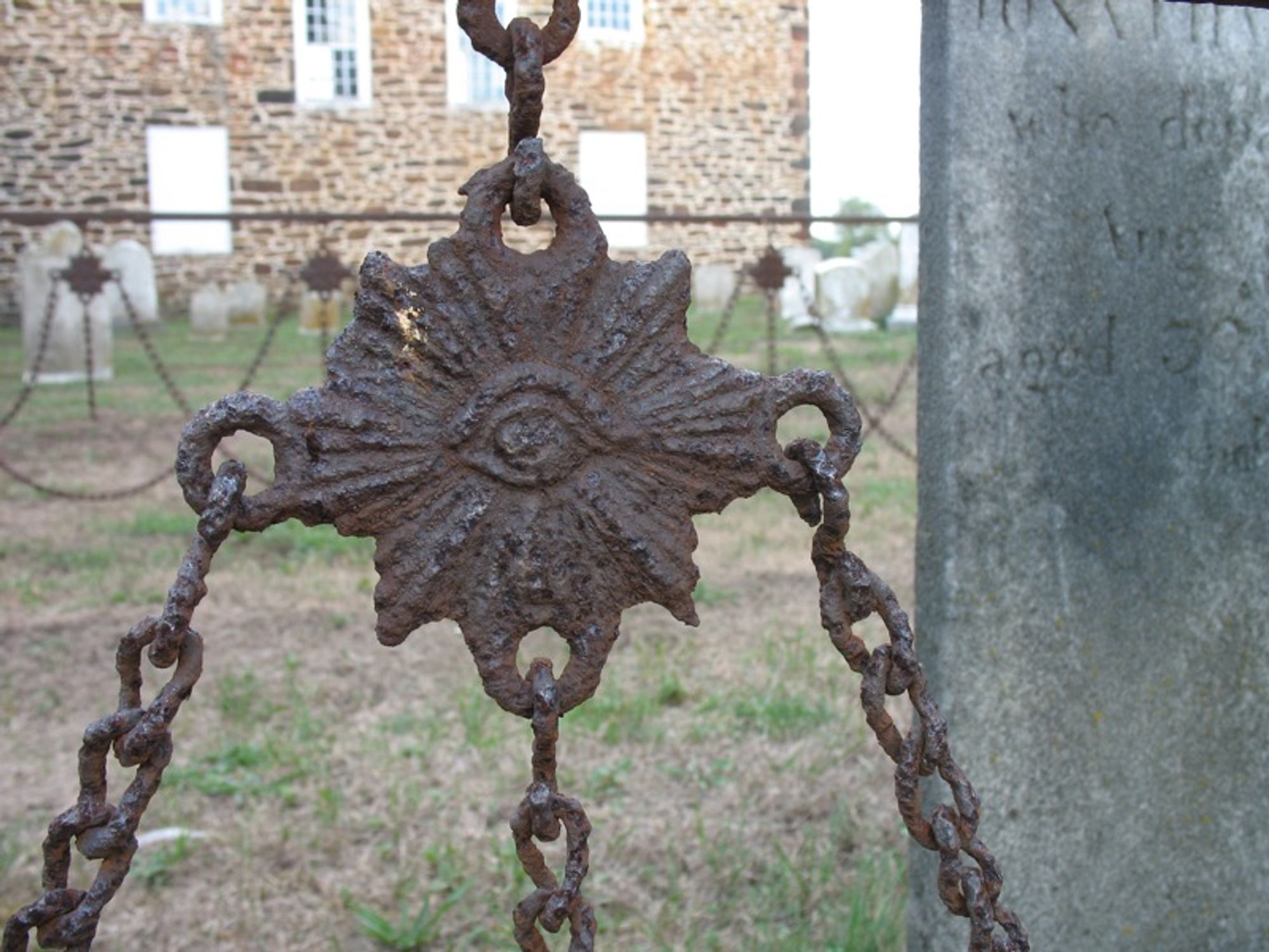 Detail of a rusty iron grave enclosure showing an eight-pointed pendant with three chains hanging from it. In the center of the pendant is an eye with rays coming from it. In the right background is the gray tombstone of Jonathan Sheppard (1748-1808). Further back are the other side of the iron enclosure, more tombstones, and the stone wall of the church.