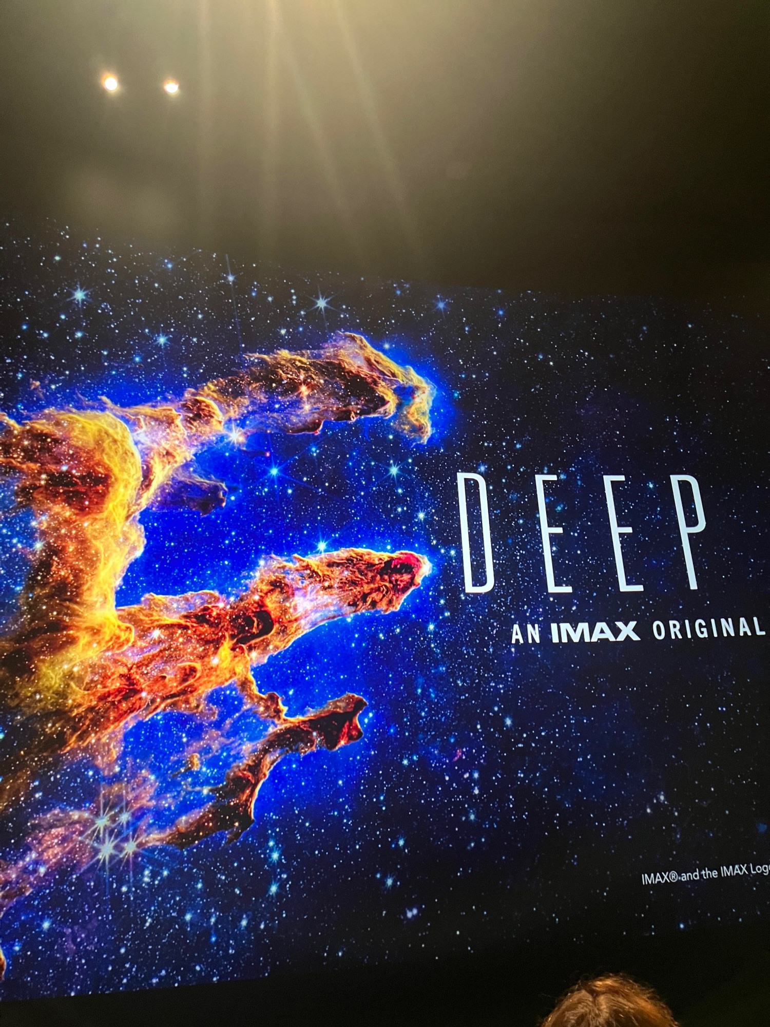 picture of an IMAX movie screen showing the text "DEEP", and below it "an IMAX original". The background picture shows orange and yellow finger like structures surrounded by blue diffuse light on a black background, with many stars in the field.