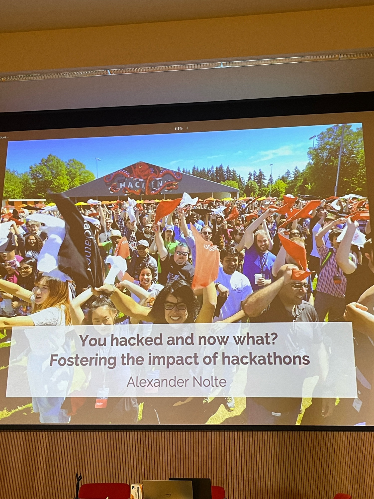 Title slide on a screen showing a happy crowd of people waving flags. The title is “you hacked and now what? Fostering the impact of hackathons”. The speaker is Alexander Nolte.