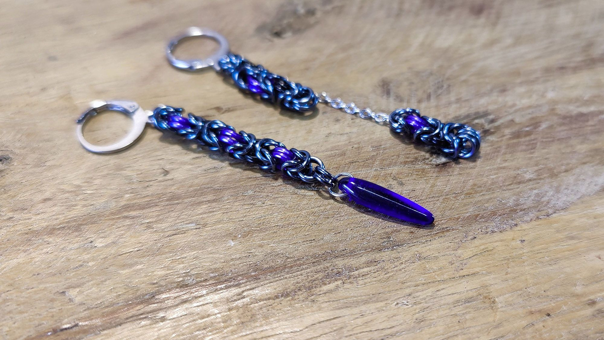 Paire de boucles d'oreilles asymétriques : l'une est une longueur de maille byzantine assez longue avec une perle en forme de dague au bout, et l'autre est comme coupée en deux avec une partie attachée directement à l'attache, et l'autre au bout d'une petite longueur de chaînette fine.