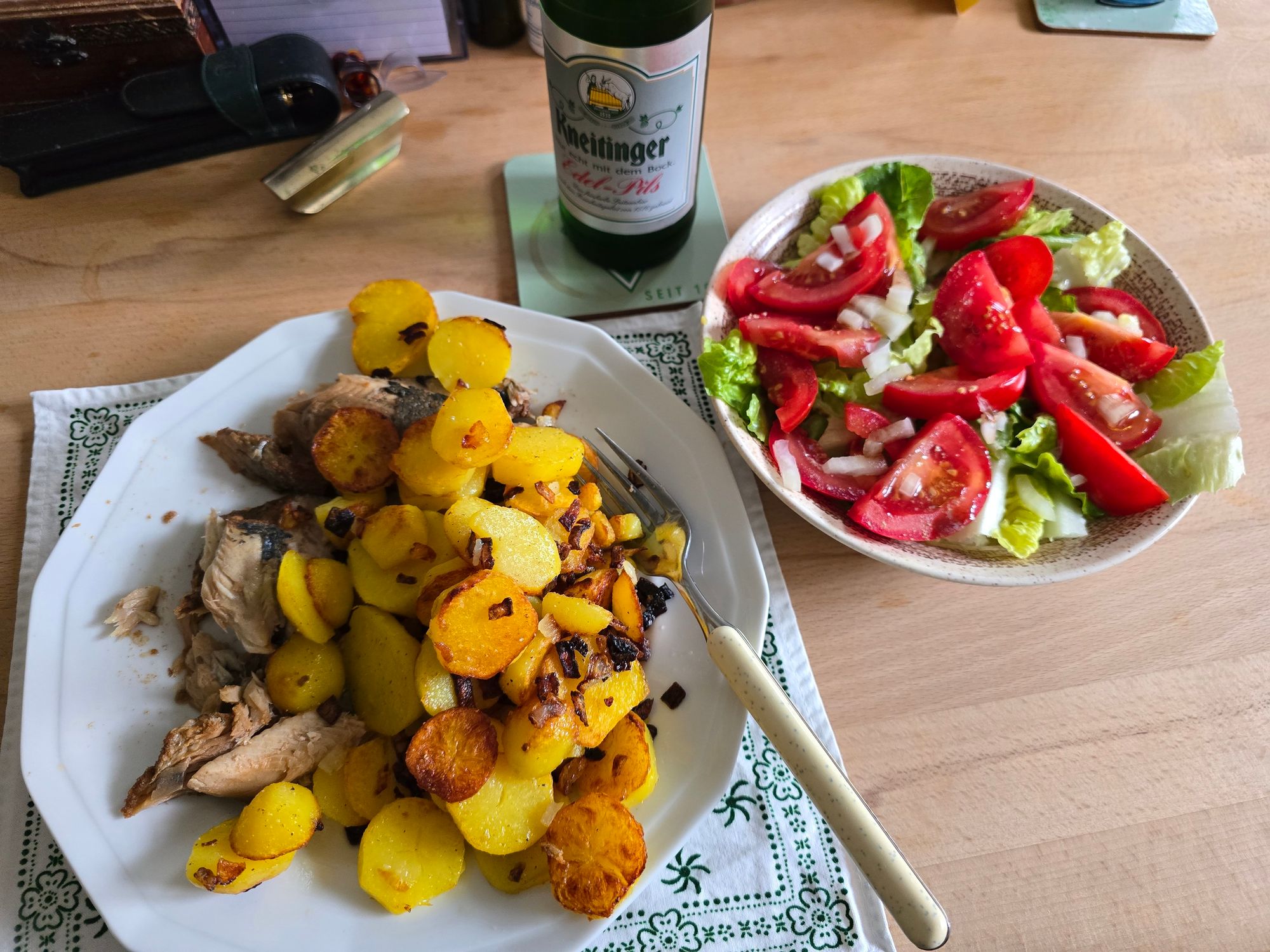 Ein Teller mit Bratkartoffeln und Bratfisch. Daneben eine kleine Schüssel mit einfachem Salat. Dabei steht ein Bier. Marke des lokalen Bieres: Kneitinger. 