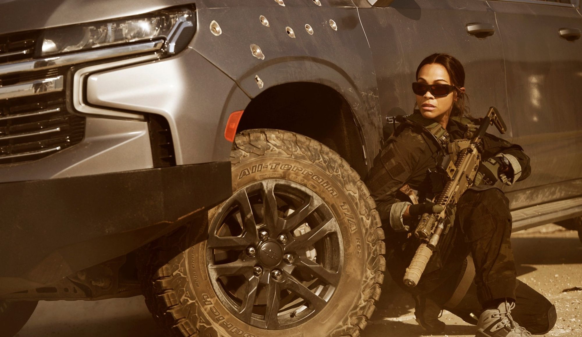 Zoe Saldana       wearing a First Spear plate carrier and SKD PIG gloves, and clutching a Sig Sauer MXC Spear with a Sig SLX forward flux silencer and an Aimpoint T2 RDS on a tanodized night vision height Unity Tactical Fast riser         crouches behind a bullet riddled SUV