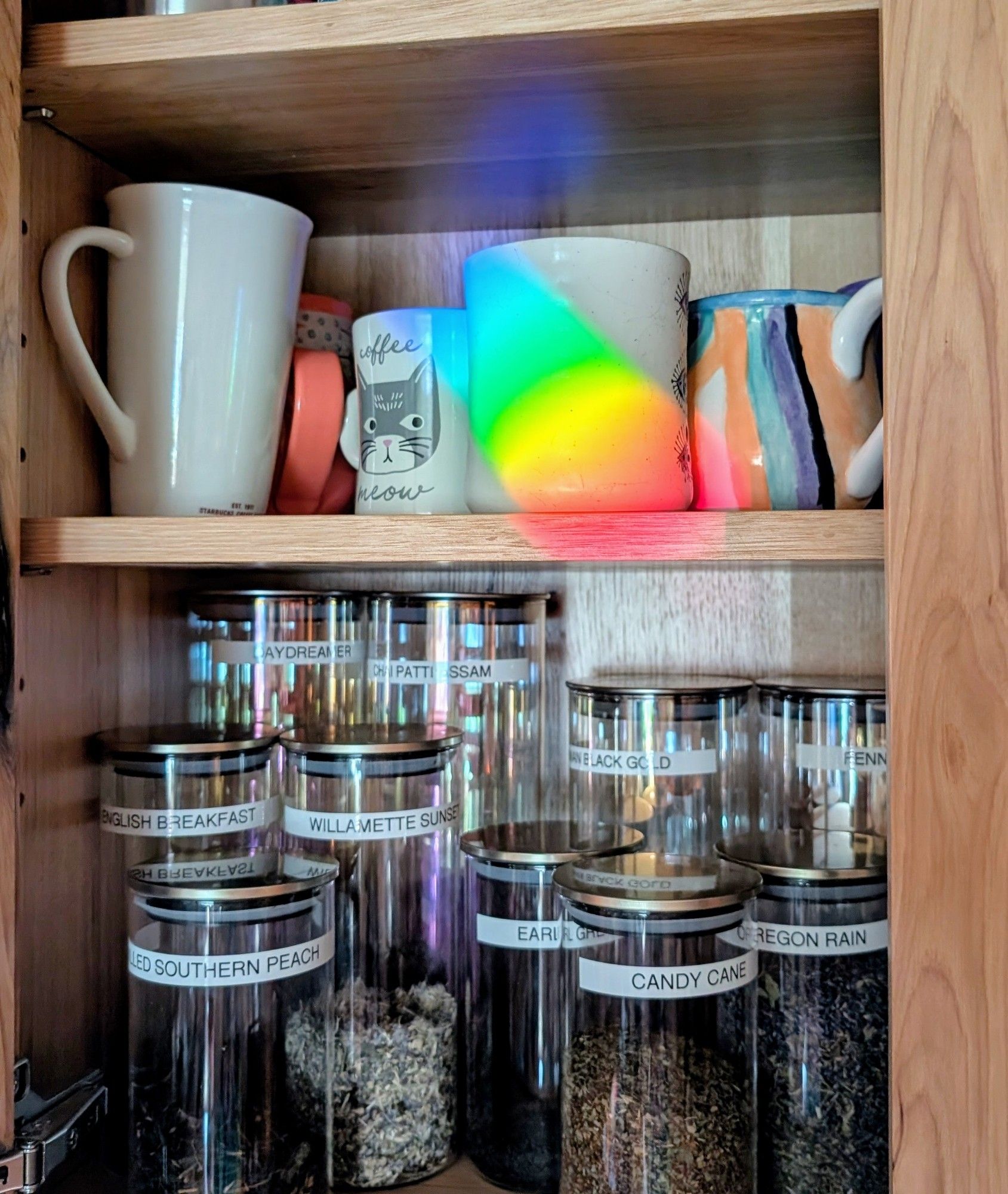 Photo of a tea and coffee cupboard. On the bottom shelf several varieties of tea are in labeled glass containers. On the upper shelf are a variety of mugs. One has a very vivid rainbow shining on it from our hanging prism in the window.