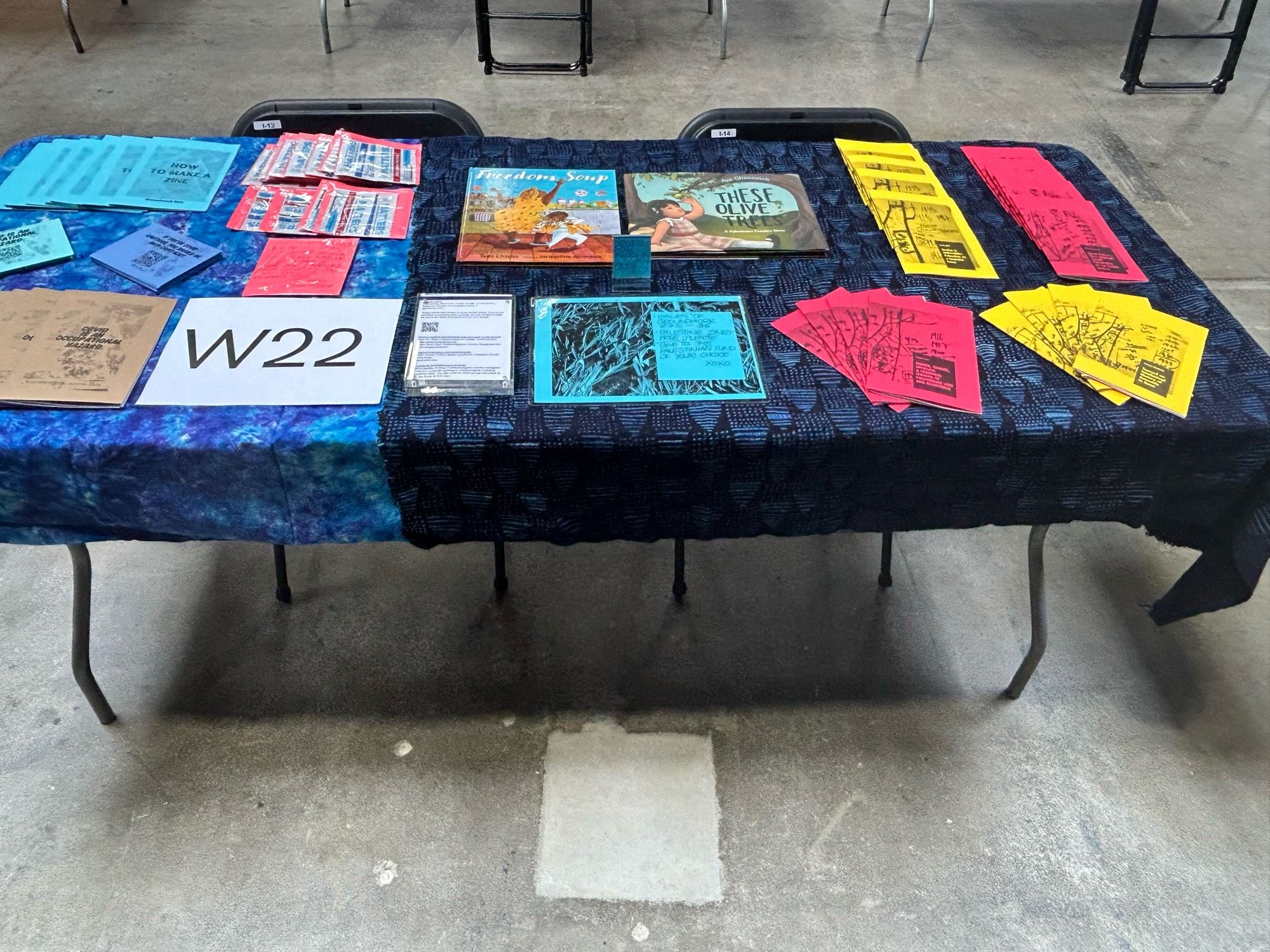 Rectangular table with blue tie dye and indigo tablecloths, table # W22, covered with zines