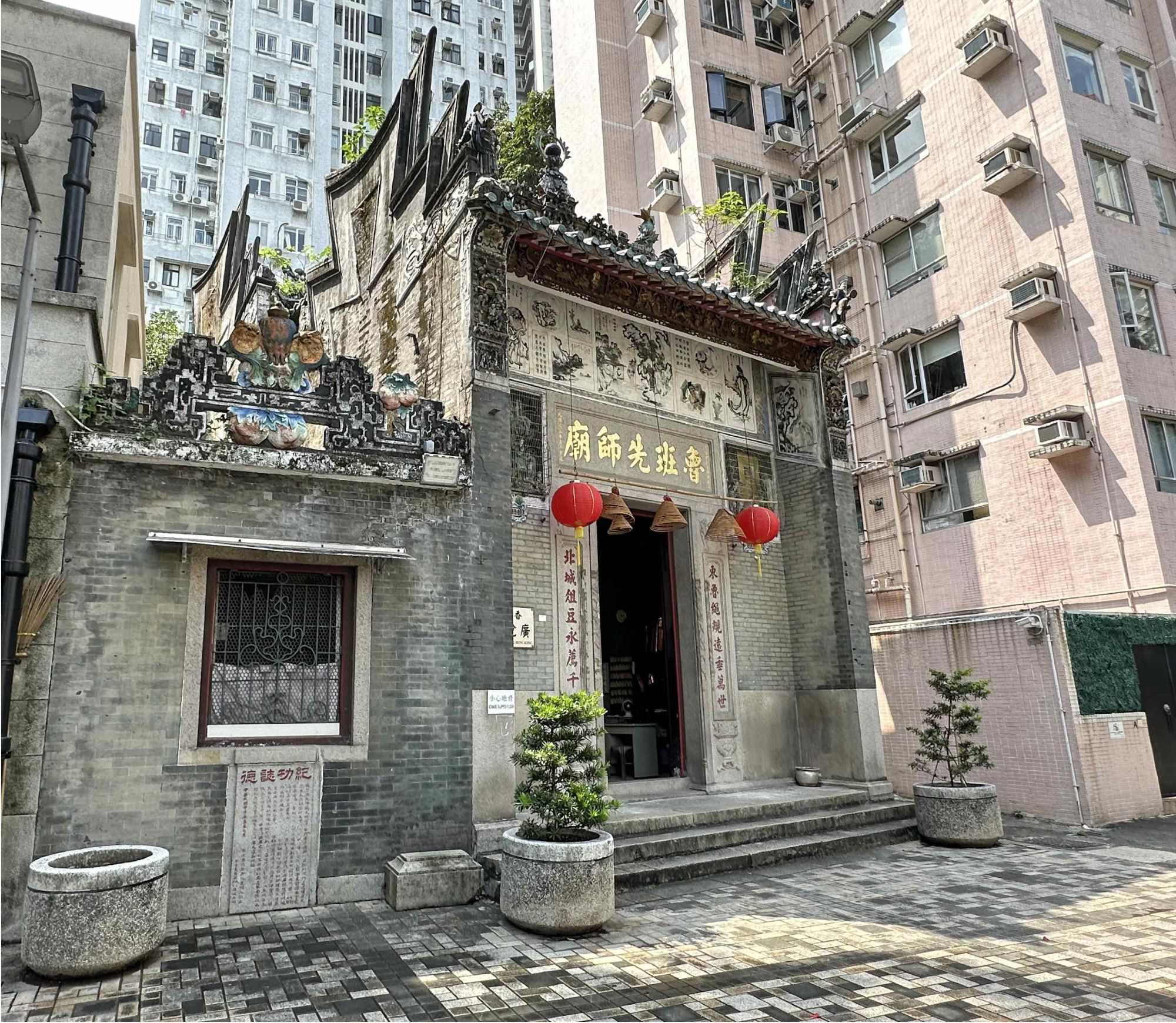 (GovHK caption) The Government gazetted today (October 10) the declaration of Lo Pan Temple in Kennedy Town as a monument under the Antiquities and Monuments Ordinance. Photo shows the front façade of the temple.

(Address) 15 Ching Lin Terrace, Kennedy Town

(Descriptive text from ChatGPT) The image shows the exterior of an old Chinese temple, situated among modern residential buildings in Hong Kong. The temple features traditional architectural elements, such as a grey brick façade, ornate decorative carvings, and a tiled roof with upward-curving eaves. Two red lanterns hang above the entrance, which is framed by stone pillars and decorated with gold Chinese characters. Potted plants are placed symmetrically on either side of the entrance, and a small stone plaque with inscriptions stands in front. The surrounding high-rise buildings provide a striking contrast to the historical and cultural aesthetic of the temple. (Reviewed by @kjoules)