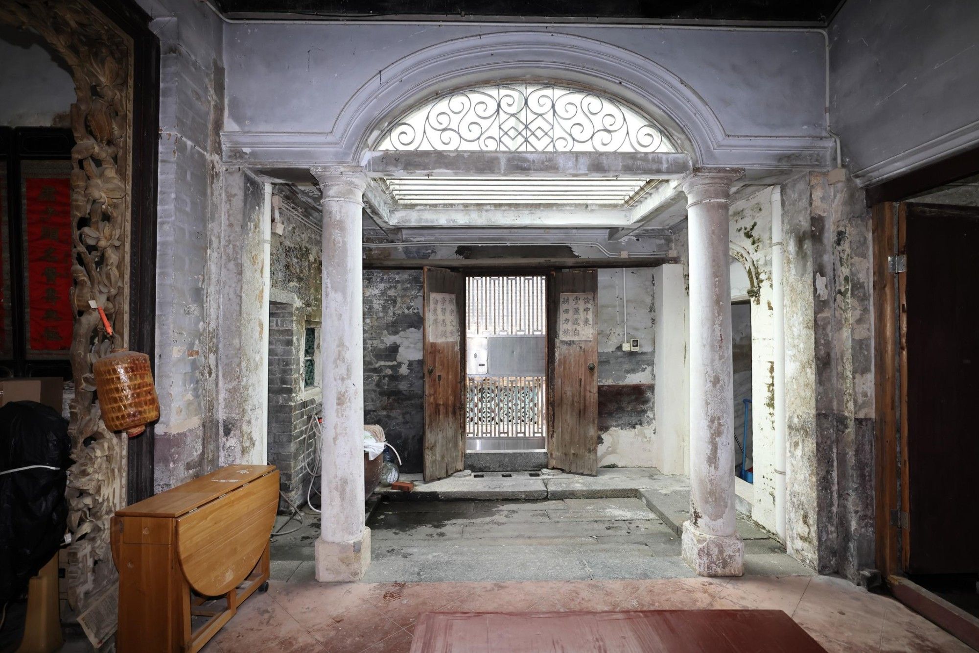 (GovHK caption) The Government gazetted today (October 10) the declaration of the Residence of Tang Pak Kau in Kam Tin, Yuen Long, as a monument under the Antiquities and Monuments Ordinance. Photo shows the light well behind the main entrance of the residence with Tuscan columns and arch.

(Descriptive text from ChatGPT) The image shows the interior of an old, dilapidated building featuring a weathered, classical architectural style. The space includes two white columns supporting an archway with decorative ironwork above. The walls and ceiling are visibly aged, with patches of peeling paint and exposed brickwork, revealing the structure’s historic nature. The floor is rough, and a small wooden table is placed in the foreground. At the back of the room, there are double wooden doors, partially open, leading outside. The scene suggests a neglected yet historically significant structure, possibly awaiting restoration. (Reviewed by @kjoules)

(Google Maps search) 錦田祠塘村20號鄧伯裘故居