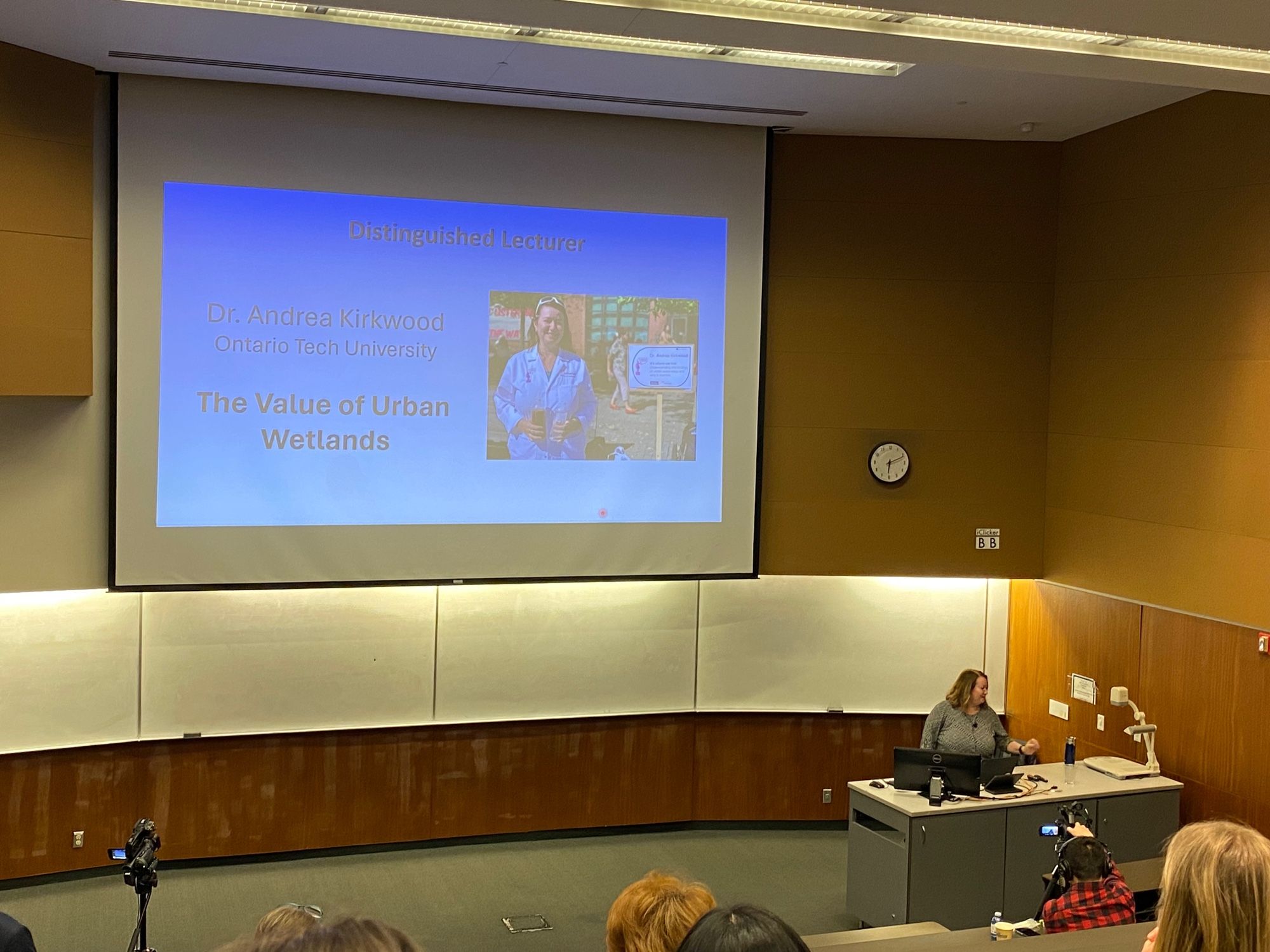 Dr Kirkwood at the podium in a large lecture hall screen shows title slide of her talk on urban wetlands