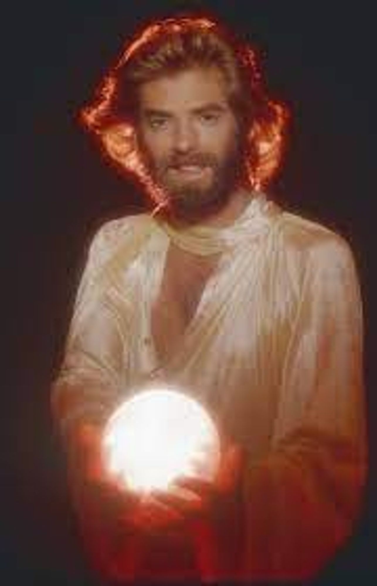 1980s Kenny Loggins pictured with flowing backlit hair, a silky flowing tunic top, and a glowing orb. He's smiling and possibly wind-blown.