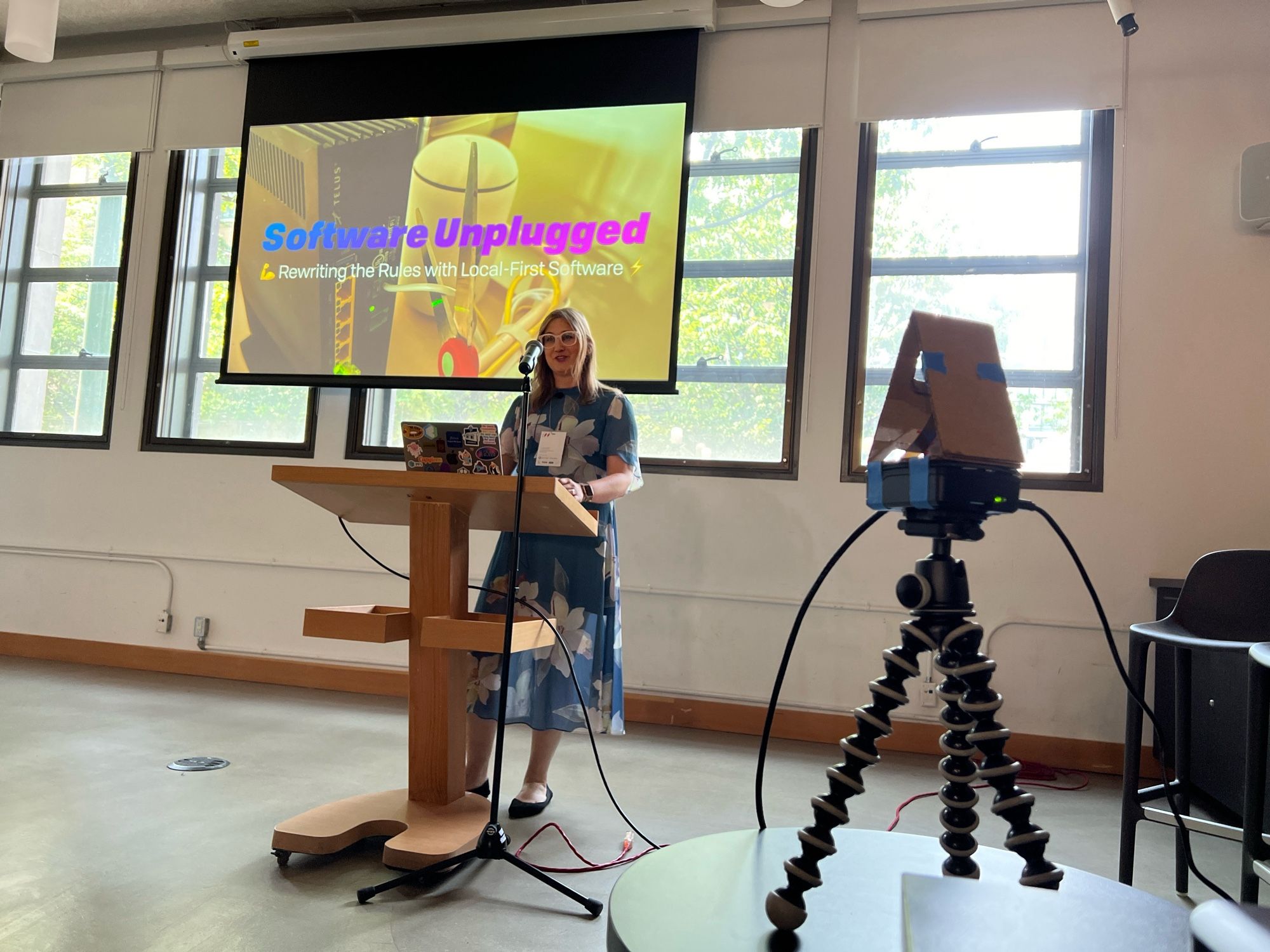 Brooke stands in front of a slide titles “software unplugged”. In the foreground is a small camera on a tripod