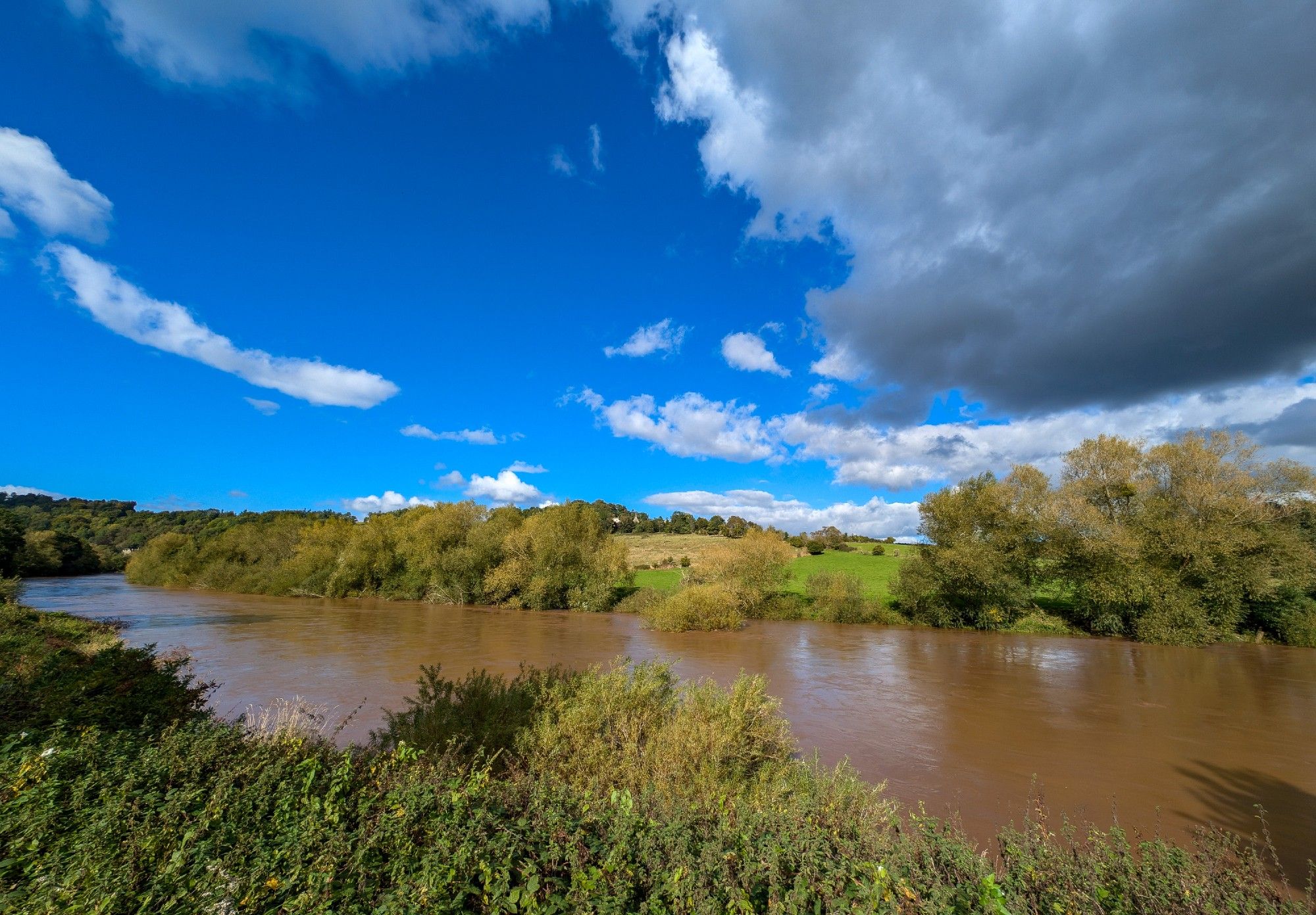 A wide angle shot of the River Wye in full spate after rainfall, with a sky of blue with some threat from the clouds.