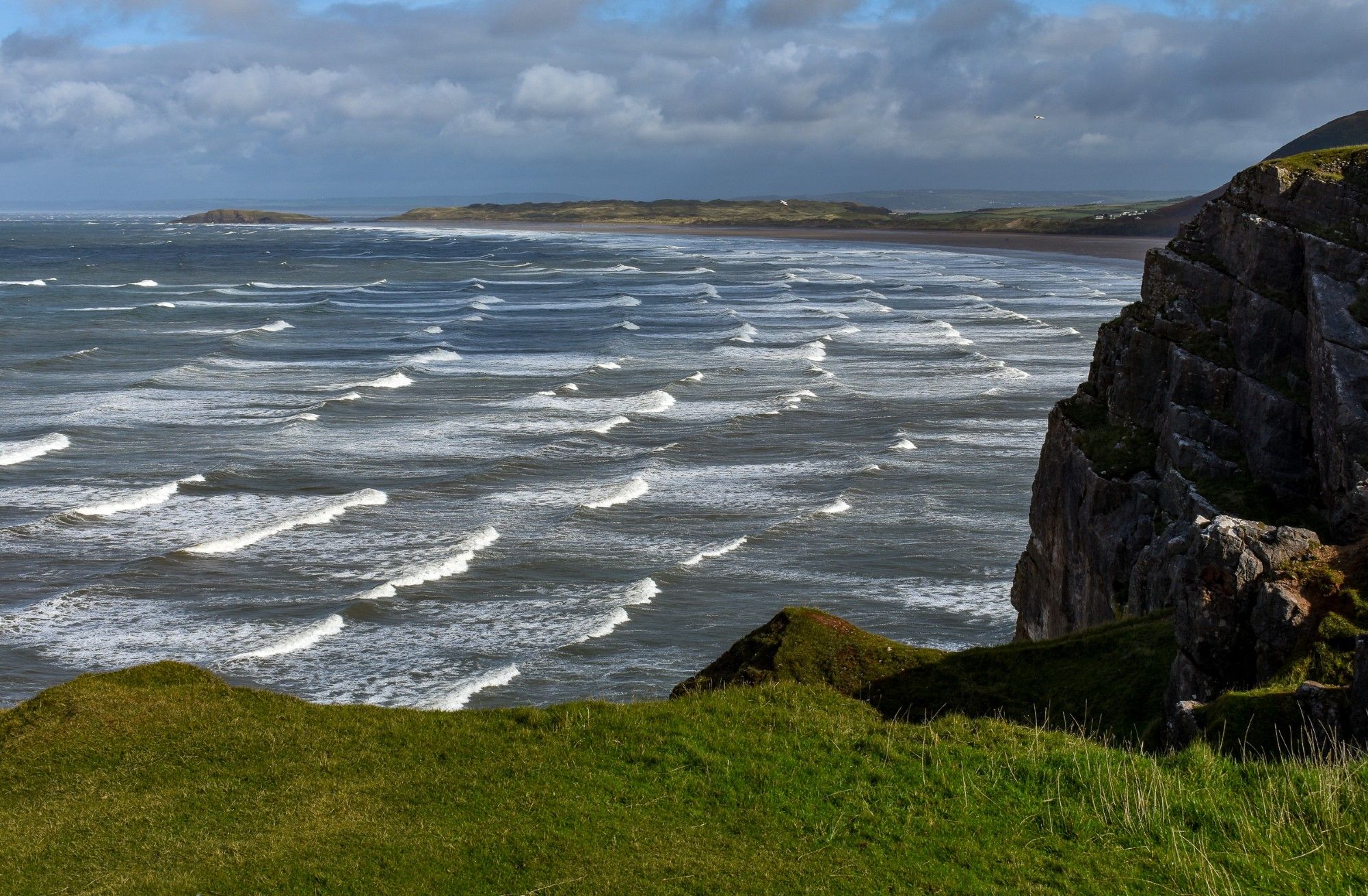 Serried ranks of small waves flow with white horses at their crests into the wide shallow bay under a September sky of blue with grey clouds.