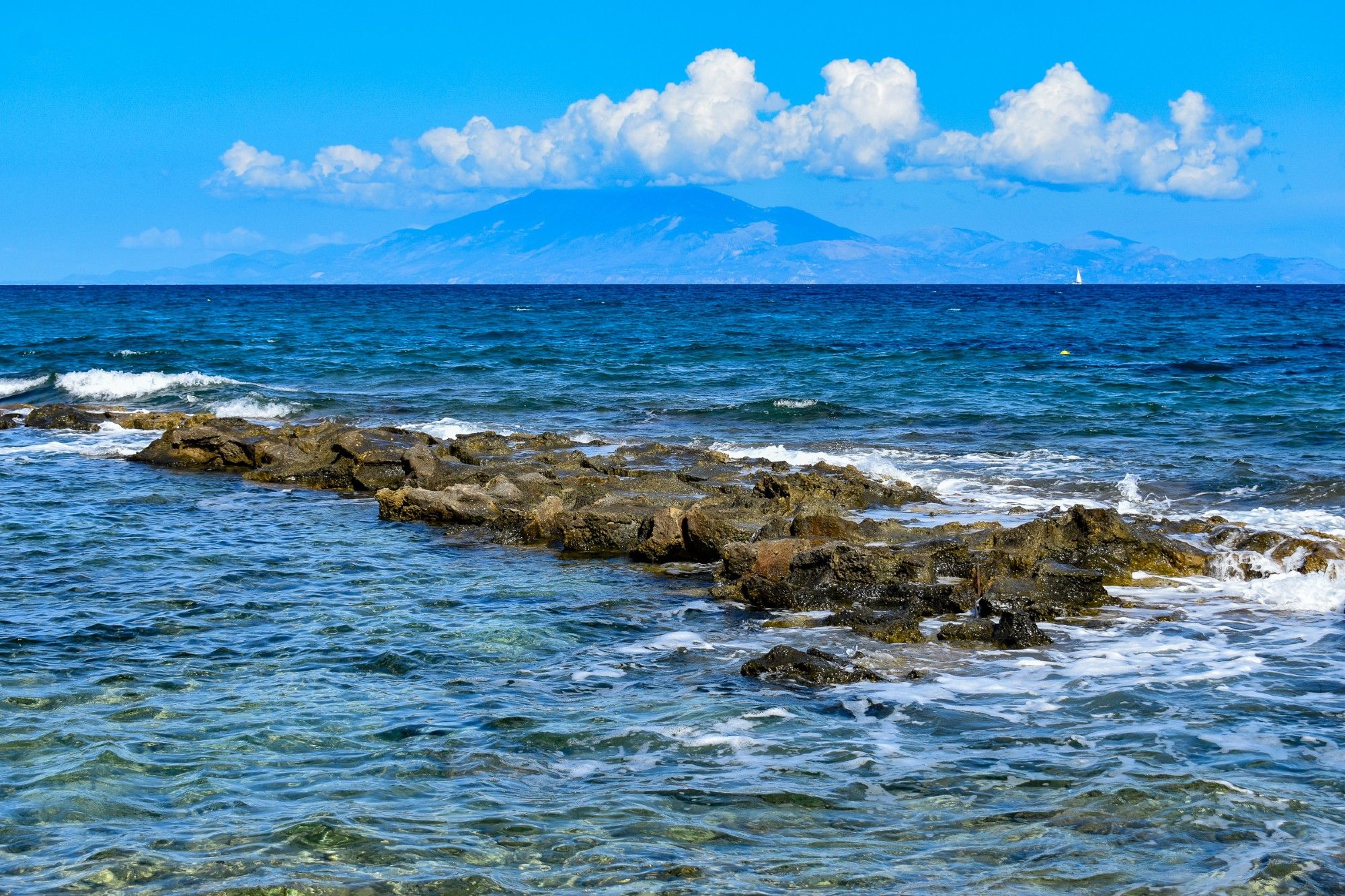 A diagonal of rocks breaks the surf of the blue Mediterranean sea on the coast of the Greek island of Zakynthos.