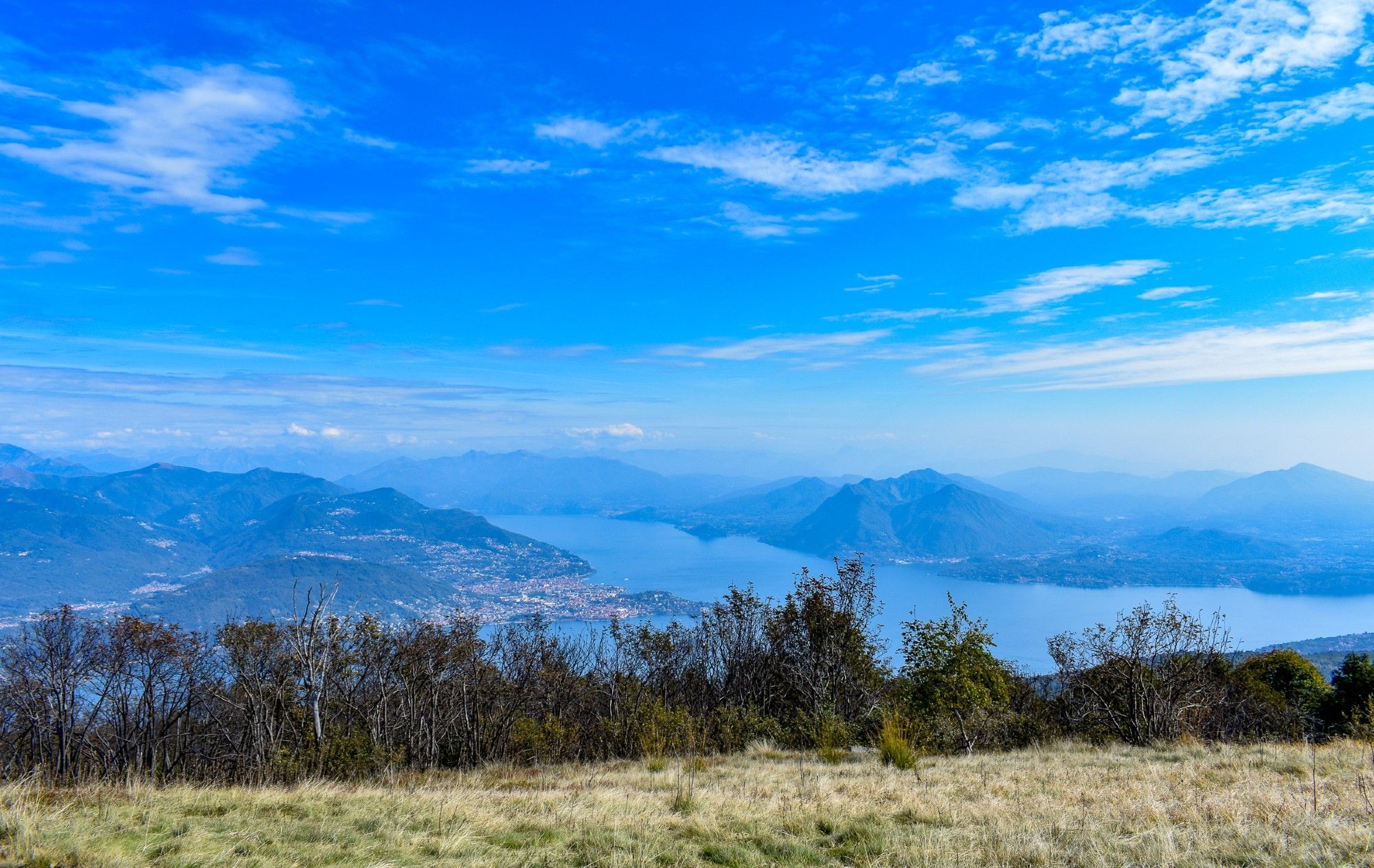 Distant mountains surround a large lake under a blue September sky patched with thin clouds. In the foreground small trees and bushes line the edge of the green field from which the photograph was taken.