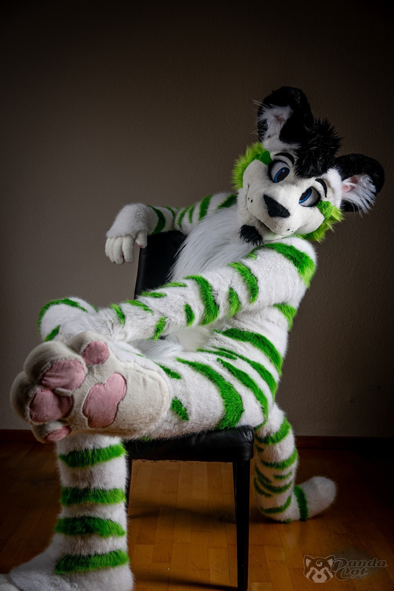 A form fitting white tiger fursuit with green stripes sits semi-sideways on a chair, facing left while turning to look directly at the camera. One arm is resting on the back of the chair, and one leg is propped up onto the other, giving the camera a good view of the underside of his white paw and pink pawpads.