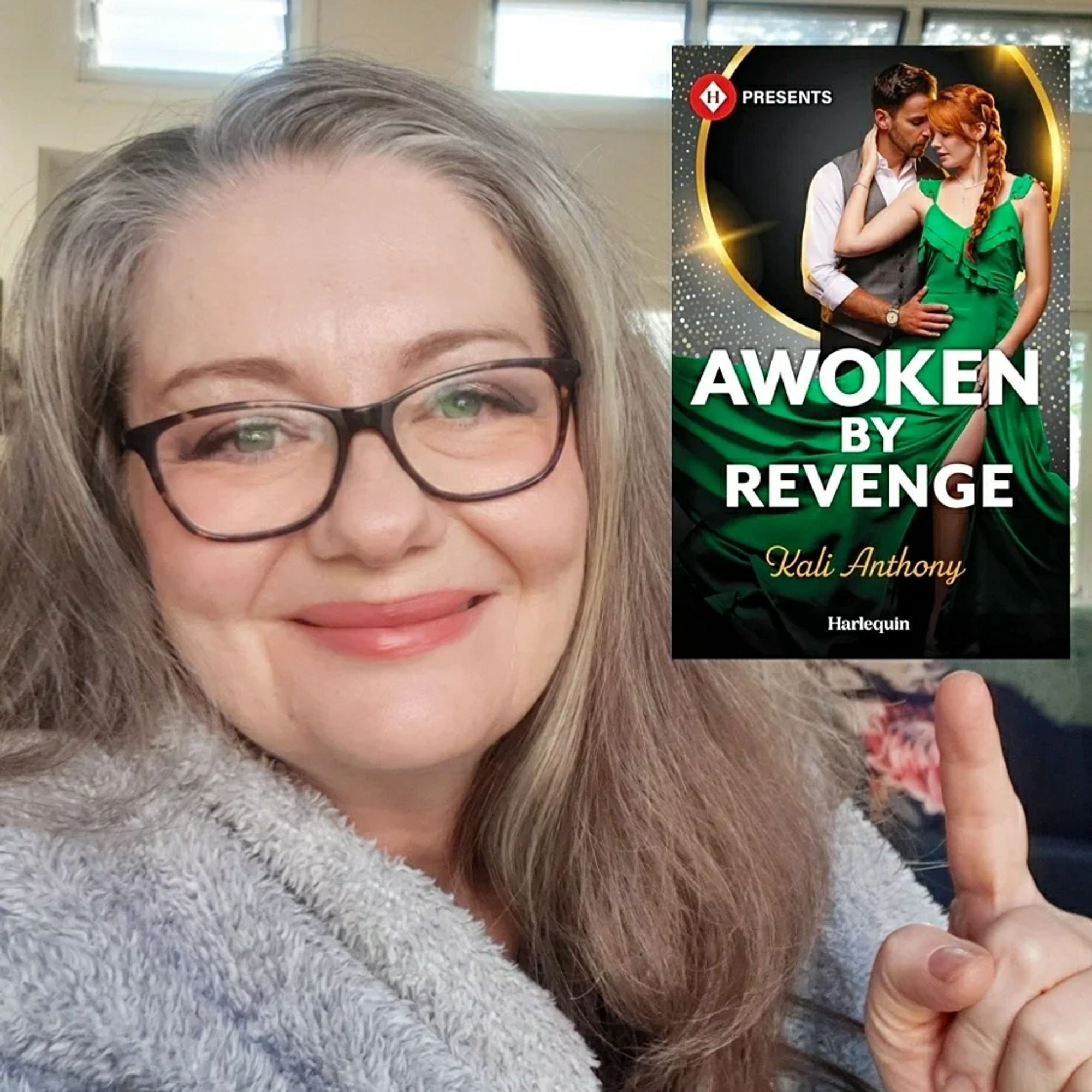 Picture of me, smiling, to the left of the photograph. I am pointing to my book awoken by revenge. The cover has a man with dark hair in a white shirt and grey vest holding a red headed woman (hair in a long plait) in a green dress.