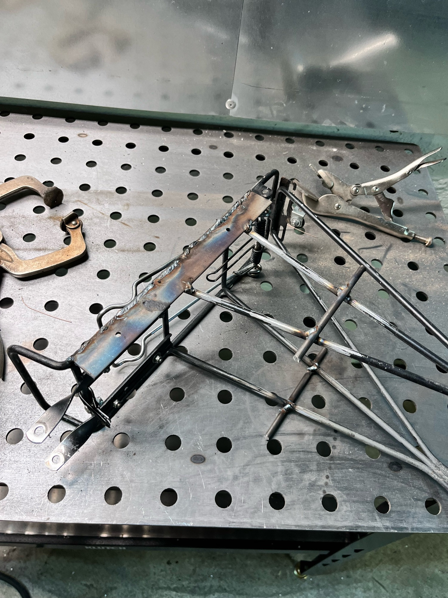 The rear rack separated from the bike on a metal welding table. The rack is unpainted, with freshly welded sections including a long square rod which will become the hard mount for the u-lock holder. The welds are rough and have not been cleaned or ground smooth yet.