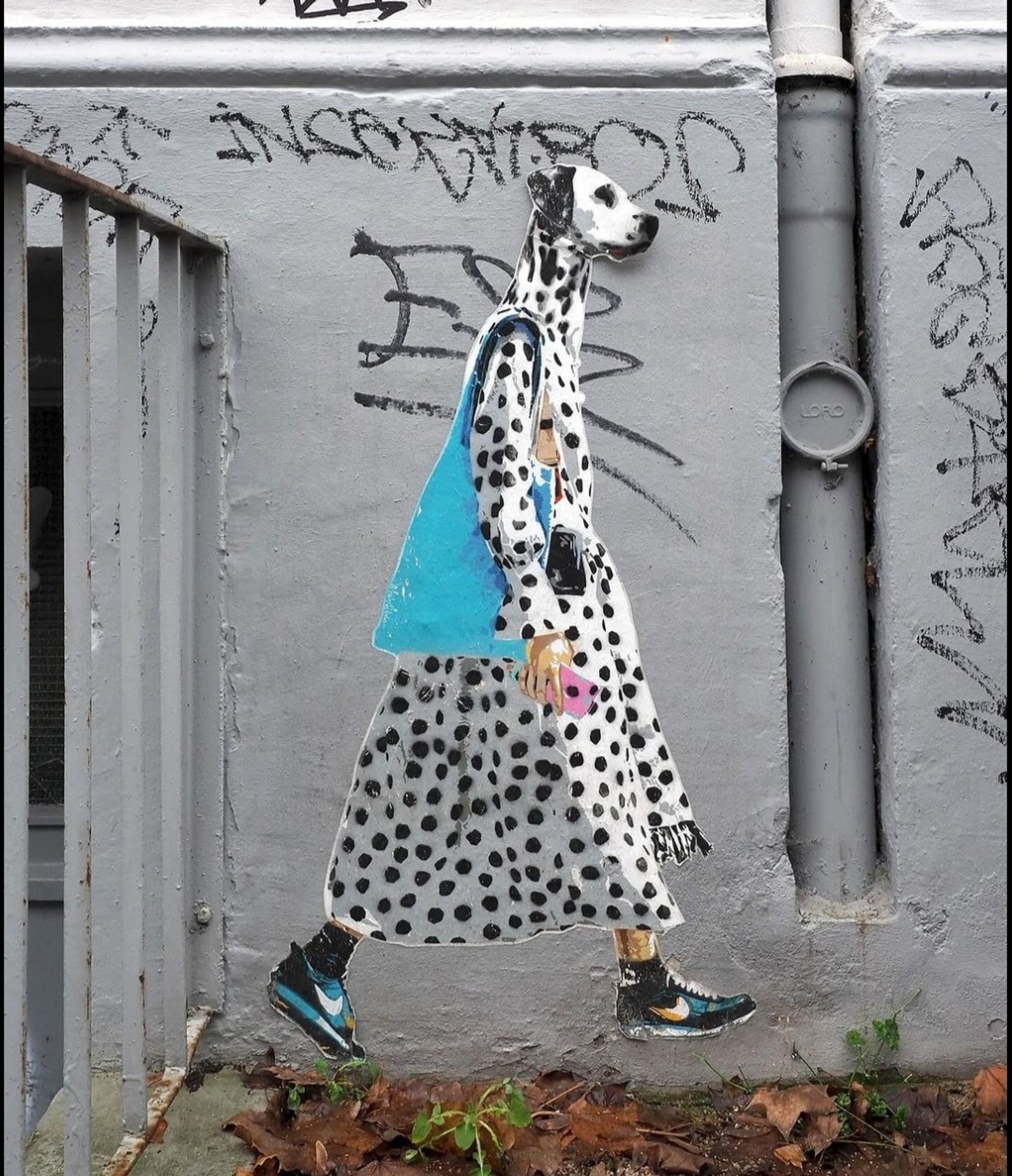 Streetartwall. On a gray painted wall with graffiti, next to a basement staircase, a paste-up with a fashionable Dalmatian dog was stuck. The small paste-up is colorful, but the upright walking Dalmatian is of course wearing mainly black and white polka-dotted clothing. A long, wide coat is combined with black Nike sports shoes with turquoise and white stripes. He also wears black socks and carries a large turquoise bag around his shoulder as an accessory. The modern dog carries a pink smartphone in his hand and strides towards spring.