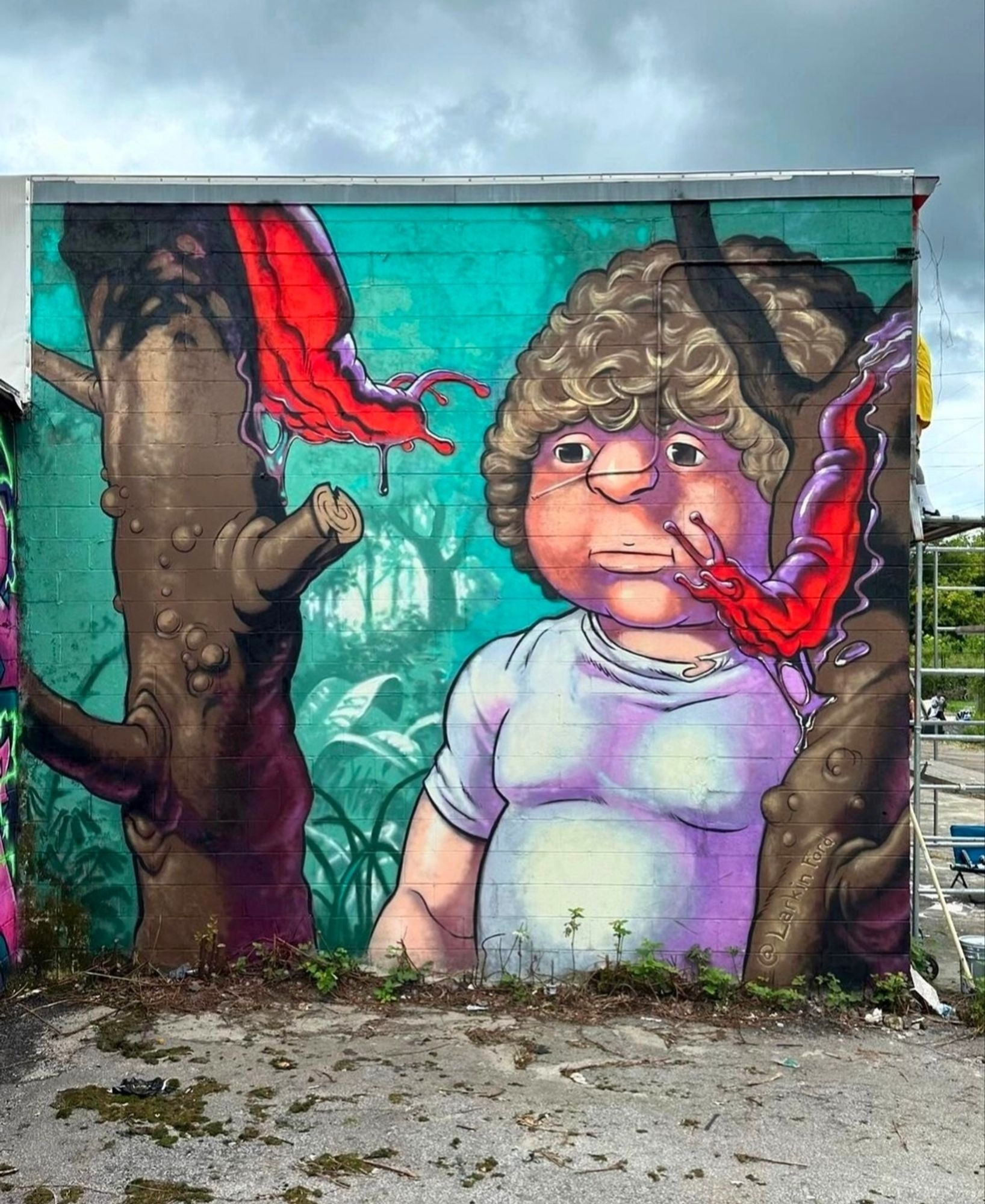 Streetartwall. The fantastic mural of a boy with two snails has been painted on an almost square outer wall of a long building. The background is a green jungle. The young man has a somewhat strange appearance. A large round head with brown curls, a thick nose, small black eyes and a rather bored expression. He is also wearing a tight white T-shirt over his not exactly slim body. To his left and right are brown tree trunks with thick red snails stuck to them. Both have turned towards him and are looking at him.
Info: The figure on the mural was inspired by an action figure from the eighties. Playskool had a series of collectible dinosaur figures. This figure was called "Zindar" and was a 3.5" tall human companion with 5 swivel joints.