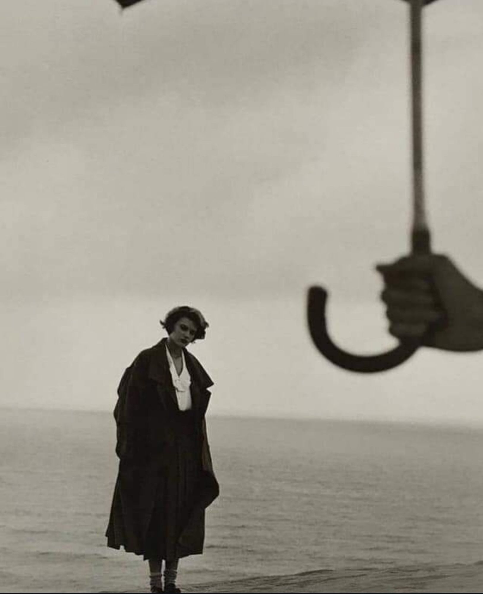 Photograph. A black and white photo of a young woman and a man in the sand dunes. The young blonde woman is wearing a long winter coat, a dark skirt and a white blouse. She is standing in front of a landscape of sand dunes that look like ocean waves. Her gaze goes to an arm in the foreground holding up an umbrella. The rather dark photo leaves different feelings and questions depending on the viewer. It doesn't seem to be raining, nor does the sun seem to be shining...
Info: Shoji Ueda (1913–2000) was a Japanese photographer who has had several gallery and museum exhibitions. One of his most popular series, is the Sand Dune series. He photographed friends and family against the background of the Tottori Sand Dunes for a poetic visual impression in monochrome. These avant-garde photos were highly praised in France, being called “Ueda Cho” (Ueda style).