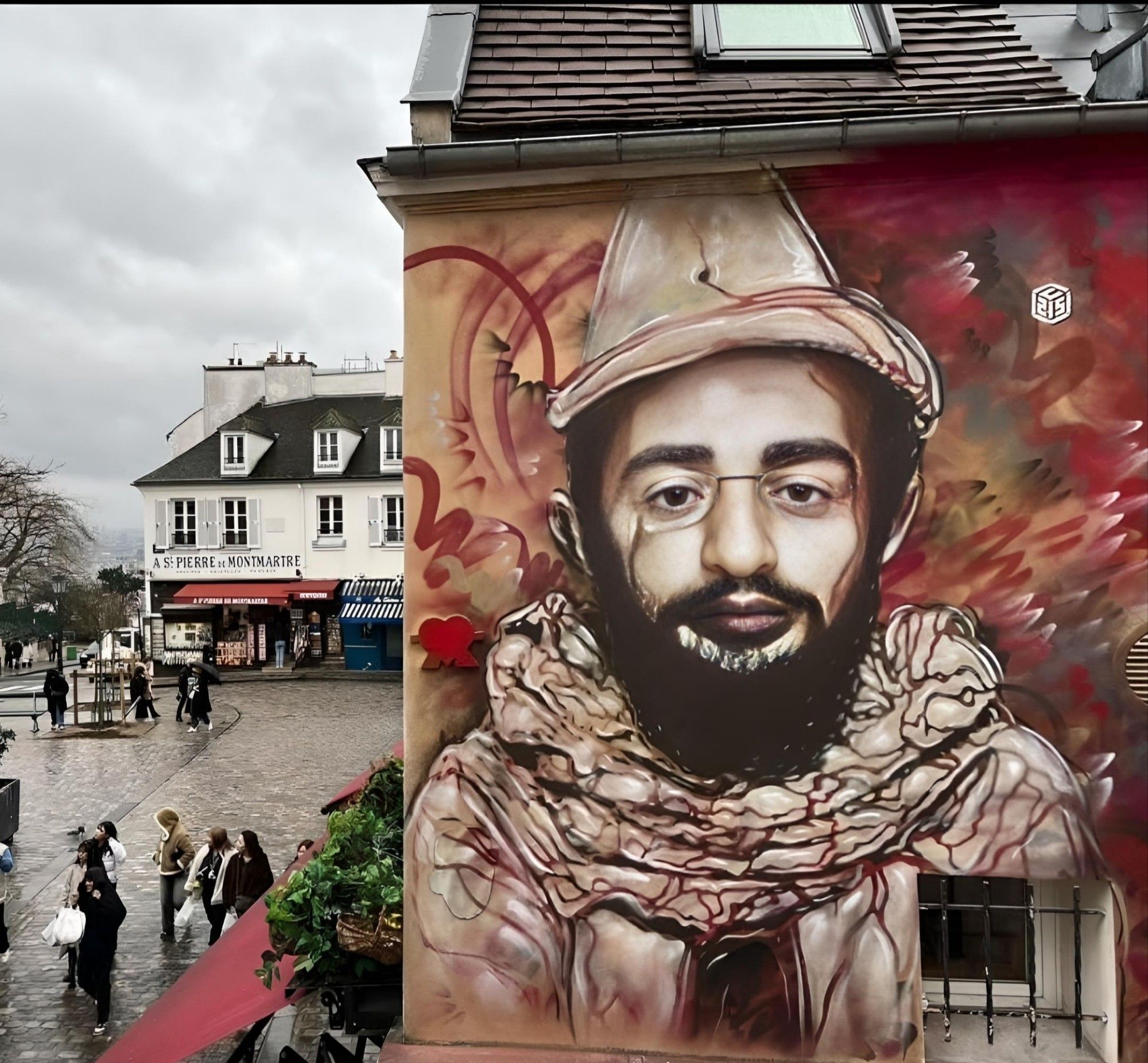 Streetartwall. A charming portrait painting was sprayed/painted on the outside wall of a two-storey building as a tribute to the painter "Henri de Toulouse Lautrec". At the bottom right-hand edge of the wall is a barred window that has been painted over. The background of the mural is in bright shades of red, with elements of yellow and orange, creating a dynamic and intense atmosphere with swirling shapes and areas of color. The portrait itself shows a man with a full black beard, broad eyebrows, small nose and small round glasses in the costume of a white clown with a matching cone-shaped foottit hat. The photo also shows the surroundings with a cobbled, busy square and buildings. A wonderful portrait of a world-famous street artist. More portraits can be seen there.
Info: There is a black and white photo of the painter Henri De Toulouse-Lautrec (1864-1901) showing him dressed as a clown. The photo from 1894 was taken as a model because it is intended to portray the painter's li