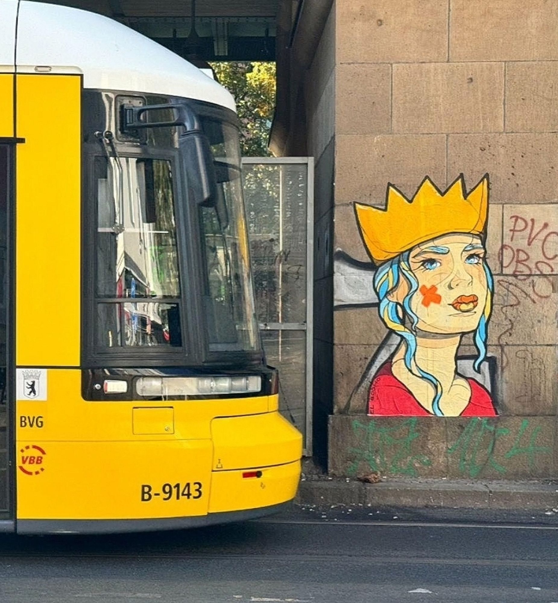 Streetartwall. The painted poster with the head of a girl in comic style is stuck to a brown exterior wall. She has long gray-blue hair and eyebrows, a beautifully drawn face and is wearing a red T-shirt. She wears a yellow crown on her head and has two orange plasters in the shape of a star on her cheek. She is looking at us. (In the photo, a yellow street train can be seen next to the mural)
Info: The anonymous street artist El Bocho is known worldwide for his self-painted portraits of women in pop art style. His art can be seen in small format as paste-ups, as posters or as large murals on walls.