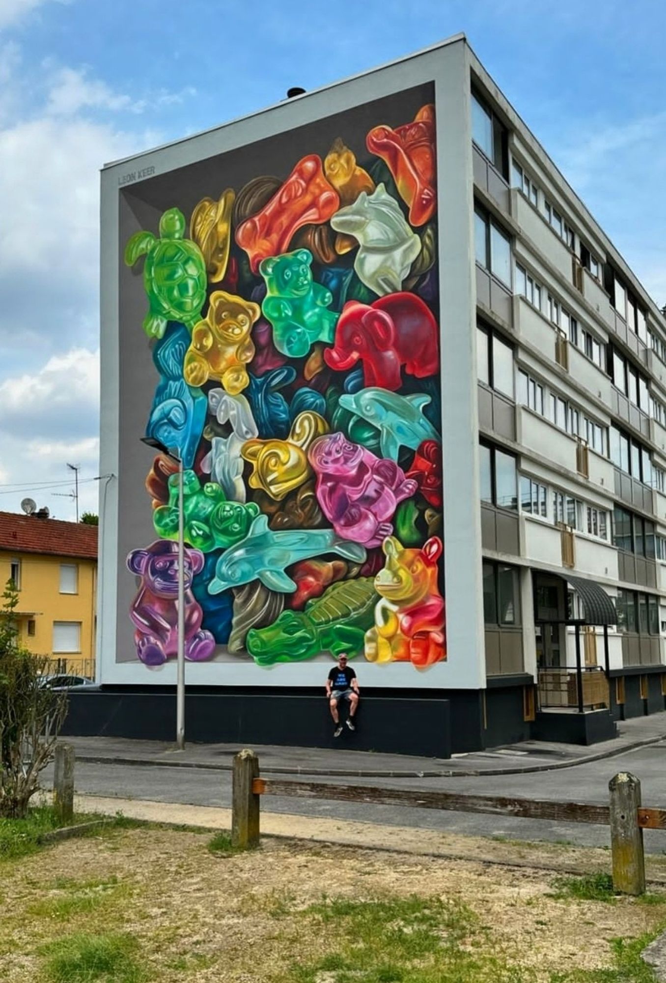 Streetartwall. A great mural with animals in 3D optics was sprayed/painted on the exterior wall of a three-story modern apartment building. The animals cavort on the wall and look like sweets in a large candy box. However, the painting shows endangered animal species, which are depicted as colorful, semi-transparent candies and thus the mural actually has a serious background. There are many animal species such as rhinos, bears, turtles, dolphins and many more. All animals that will soon only be seen as sweets and in zoos.
(In the photo the artist is sitting in front of his mural)