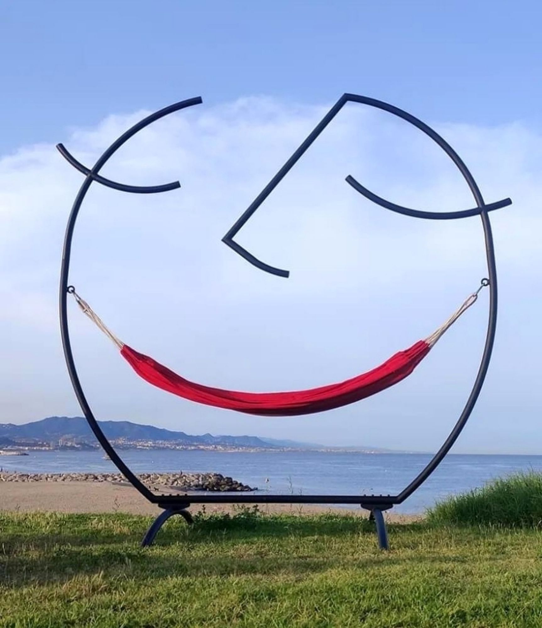 Photography. A color photo of an exceptionally cheerful sculpture. The photo shows a sculpture made of a round black metal frame (approx. 2m66 high). The metal arch is open at the top, on both sides there are two side struts bent upwards (like eyes) and an L-shaped strut (nose) extends to the top center. A red hammock (mouth) hangs in the middle. Together, the structure forms a smiling face. The unusual hammock stands on a green meadow and a blue sea with hills can be seen in the background.
Info: The happy hammock is a sculpture by architect and designer Federica Sala. The sculpture is part of a series of kinetic sculptures by the studio. The idea is to create a simple red cradle, an empathetic object that embraces the inner child in us and allows it to enjoy, relax and reflect.