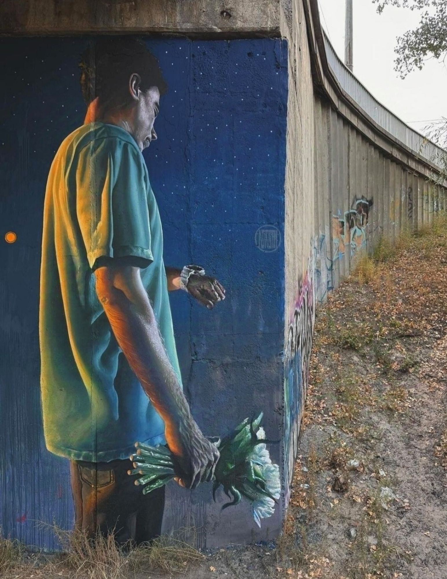 Streetartwall. A beautiful mural of a man waiting for his Girlfriend was sprayed/painted on a concrete wall under a bridge in Kiev. The mural shows the dark-haired man in a green T-shirt from behind, looking at his wristwatch. He is holding a bouquet of flowers in his other hand. Around him, the night has already turned a deep blue. The moon is full and small stars are shining. (The photo shows the long wall next to the mural, the foliage and the path leading up to the road).
Info: Sasha Korban about his Mural: 
"Yeah, there's a war in our country, but we're still going on dates, falling in love, living our lives."