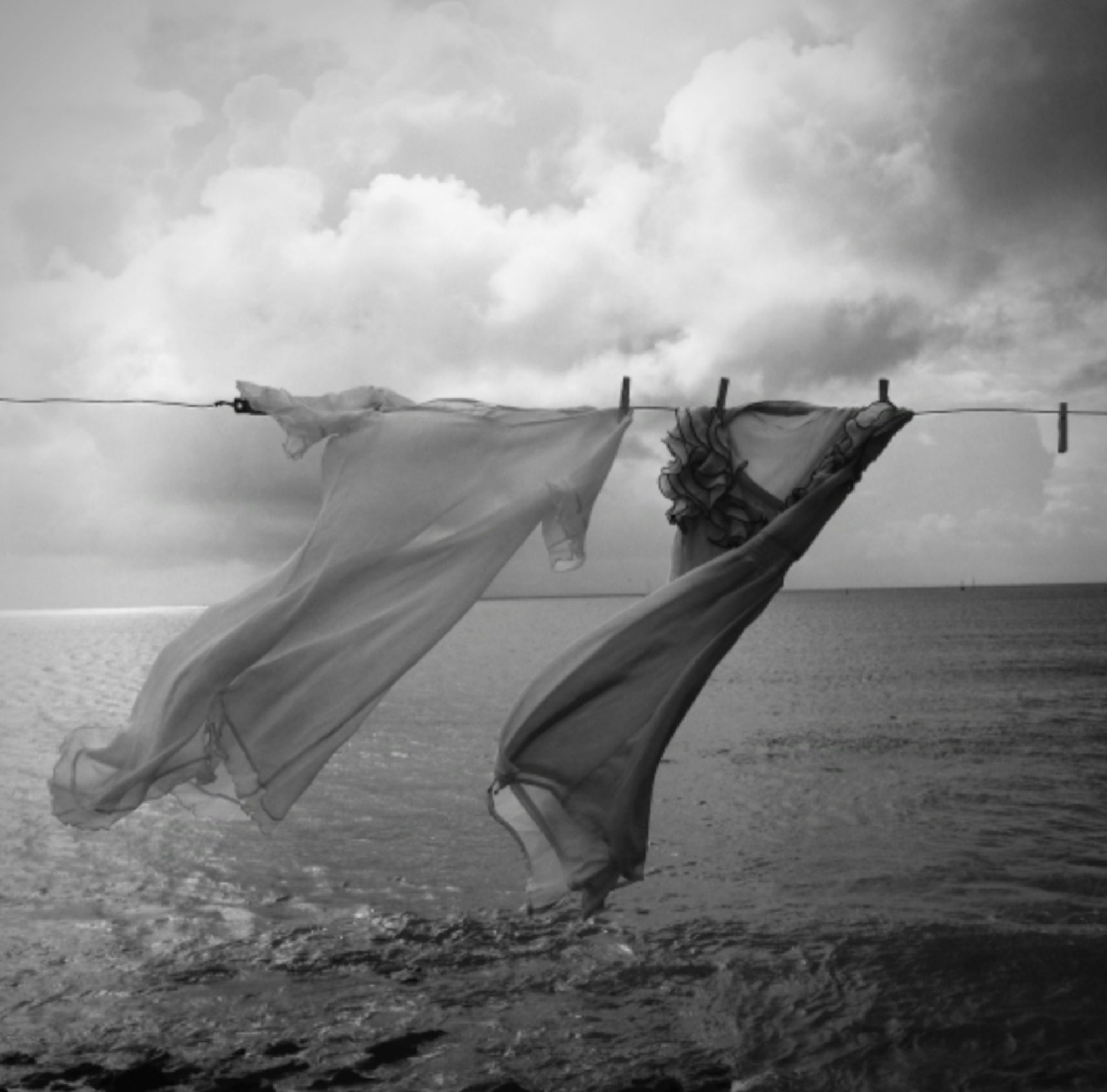 Photography. A black and white photo of two summer dresses on a washing line by the sea. The almost transparent-looking long dresses (one white and one slightly darker) flutter to the left in the wind. As if they were dancing and swaying to the rhythm. The sea can be seen in the background and a gray wall of clouds on the horizon. The photo radiates both the calm of a day by the sea and the restlessness that heralds the approaching storm and perhaps even the end of summer. When you think about this photo, you strangely remember the scene in color. The head continues to tell the story...
PhotoInfos: Taken and processed with Cameramatic app/Apple I-Phone
/2.4 /4.1 mm/Iso50