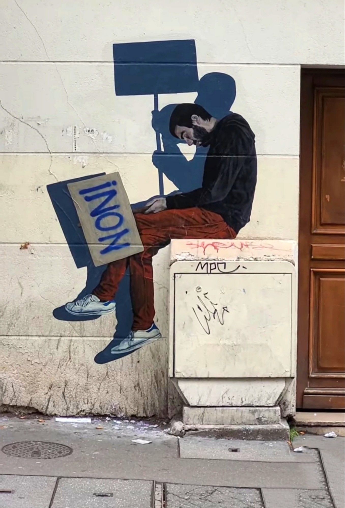 Streetartwall. The life-size, self-painted picture of a sleeping man has been stuck to a yellow house wall. The dark-haired man with a full beard sits huddled on a (real) distribution box and sleeps. He is wearing red trousers, a black sweater and sneakers. He is holding a sign with the French word "non" (English: no) written on it. Behind him is his shadow, who is not sleeping but holding up the protest sign.
Info: Notes by the artist Levalet: "In these troubled times, we can remind ourselves that each of us has the power to say no, to resist, even when the worst seems certain. Let's wake up!"