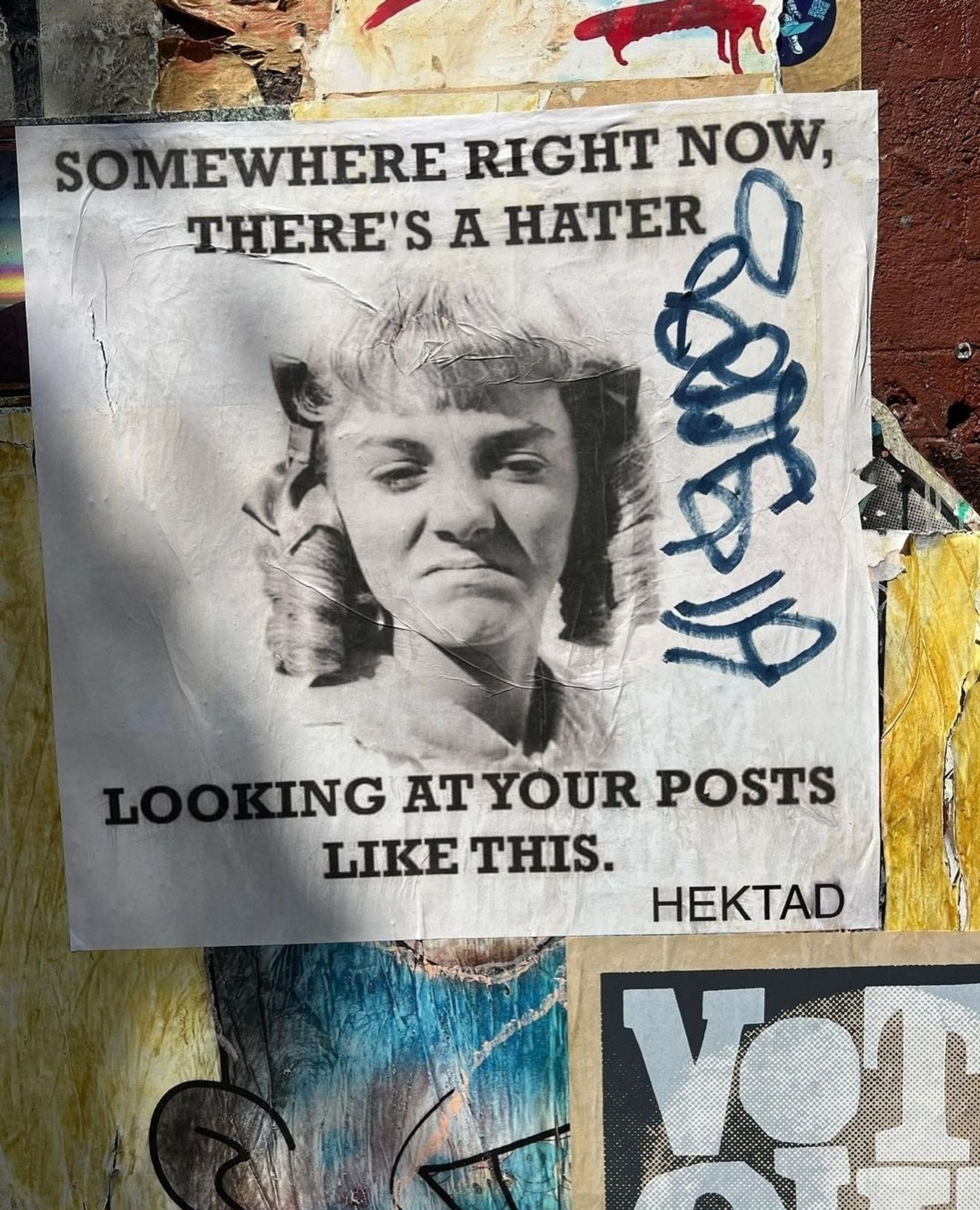 Streetartwall. On a colorful graffiti wall is a simple black and white poster that reads, "Somewhere, a Hater is sitting right now looking at your post like this." Accompanying it is the face of a blonde girl with twisted curls and short bangs, wrinkling her nose in disdain. It is the actress Alison Arngrim in her role as Nellie Oleson from the American series "Little House on the Prairie" from the seventies.