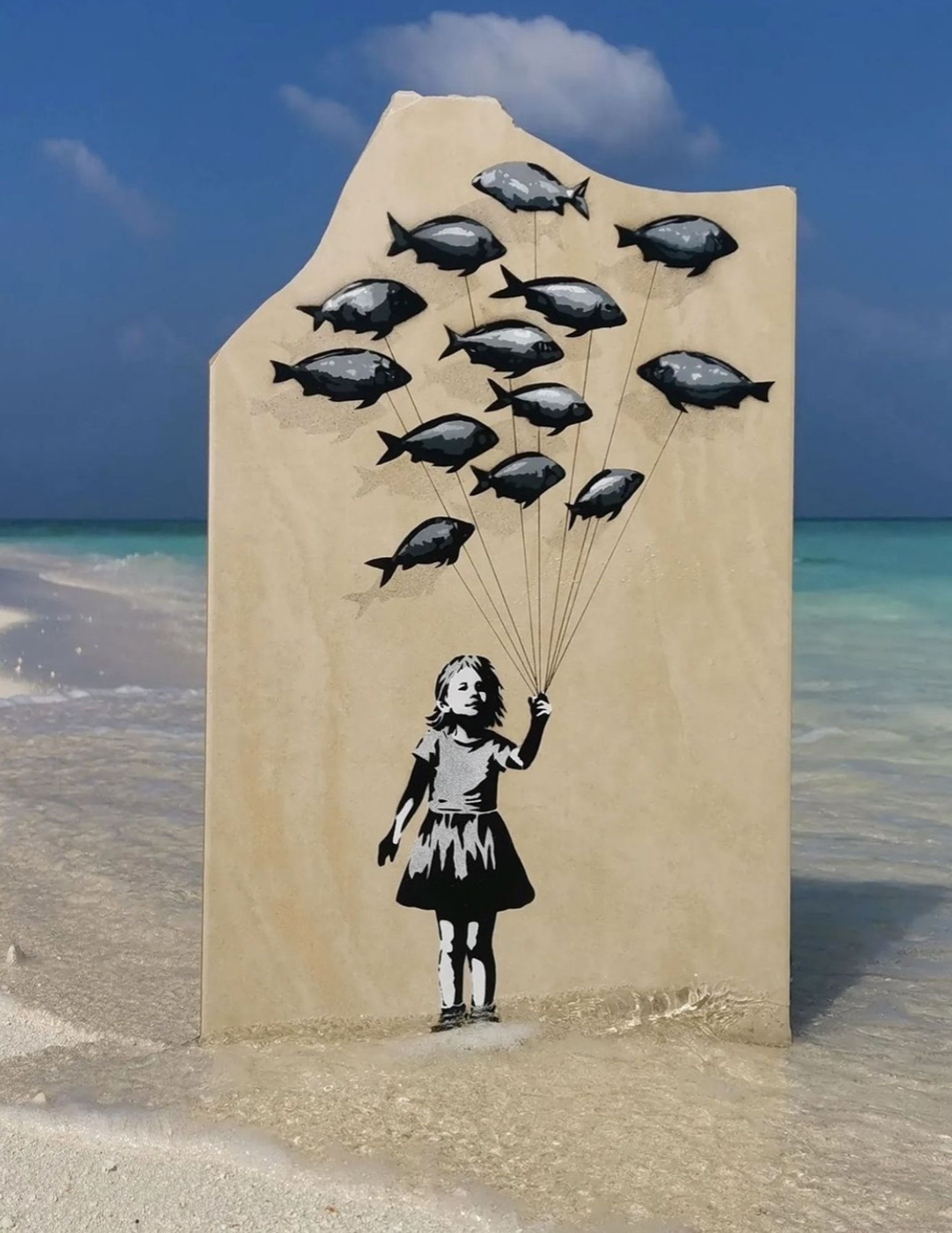 Streetartwall. A cute mural of a little girl was spray-painted on a beige life-size wooden wall, which was placed in the sea near the beach. The figures have been sprayed in shades of gray, black and white. A little girl in a short dress holds 13 small balloons floating above her. They look like little gray fish. Flying fish on the beach.