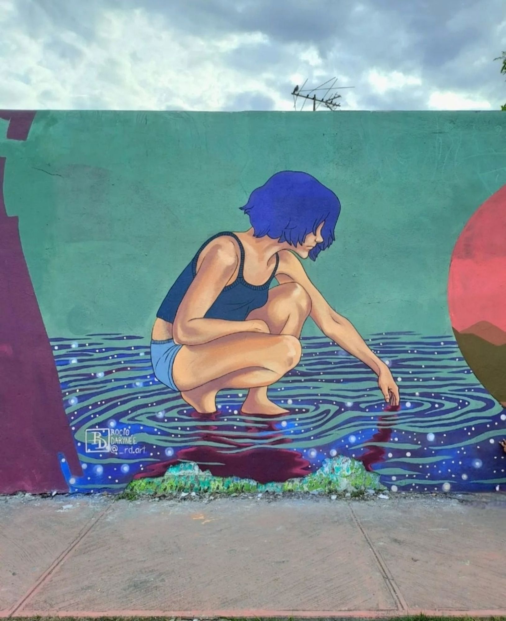 Streetartwall. An approx. 3x3 m mural with a girl by the water was sprayed/painted on a street wall in a naive painting style. The girl has a blue short hairstyle and wears a dark blue top and short light blue shorts. She is squatting in low water and holding a finger in it. The green-blue water draws circles and small glittering pearl dots shine brightly. Above her is a green background, with an implied red circle of the sun on the right and a brown rock on the left. A beautiful little imaginative mural. (The photo shows a blue-grey sky and a television aerial above it).