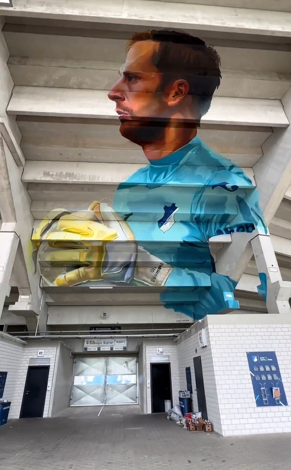 Streetartwall. A huge mural of a soccer goalkeeper with a ball has been sprayed onto the concrete ceiling under the stands of a soccer stadium. The dark-haired man with a full beard is wearing the green club shirt and goalkeeper gloves. He is holding the ball in his hand and looking to the left. 
Info: The realistically designed mural is also a tribute to goalkeeper Oliver Baumann. He currently plays for TSG 1899 Hoffenheim in the German Bundesliga, where he played his 300th Bundesliga match, for which he received this mural as a tribute.