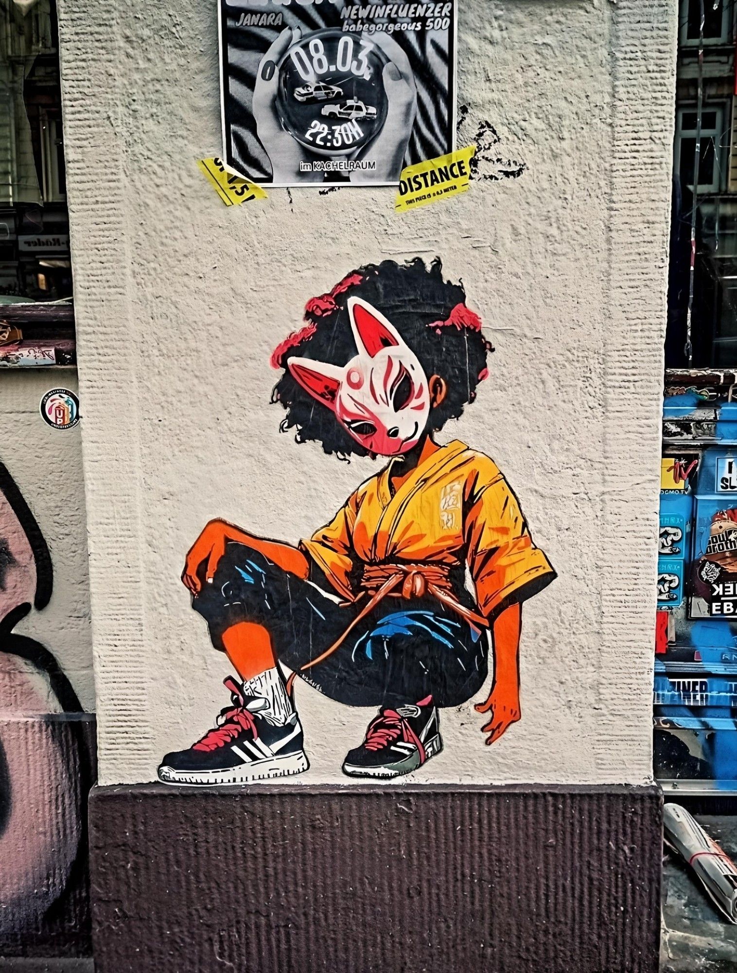 Streetartwall. A small mural of a young woman in comic style has been sprayed onto a white wall next to a shop window. The small mural has a colorful design and shows a young woman dressed in hip-hop style with blue pants, a yellow shirt and sports shoes, squatting on the ground. She is finely drawn, has wild black hair and a mask in front of her face.  The mask is reminiscent of a traditional Asian cat representation. The girl is looking in the direction of the viewer. (In the photo, an event poster hangs above her and there are flyers and graffiti to the left and right)