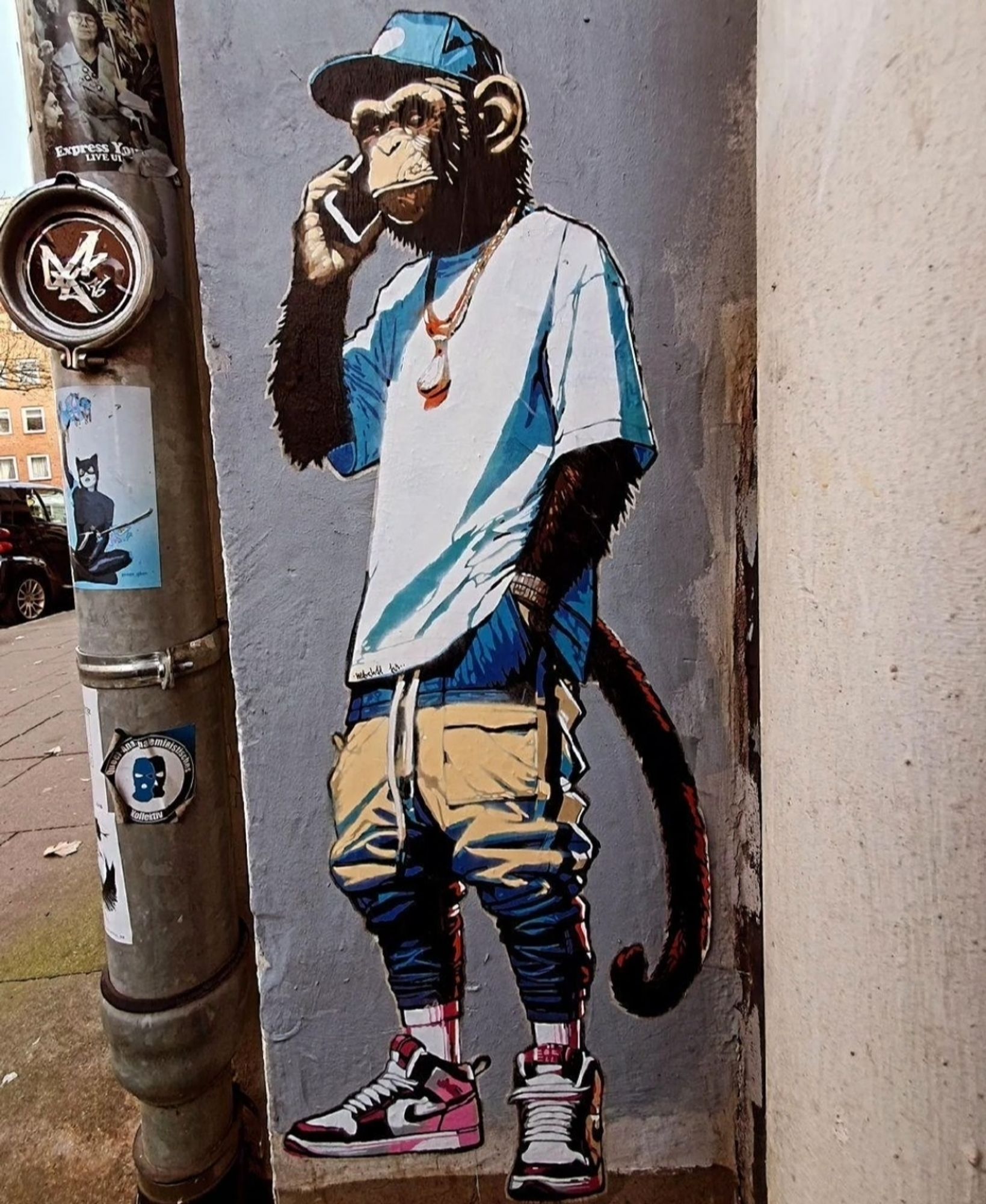 Streetartwall. A small comic-style mural showing a monkey making a phone call was sprayed onto a narrow gray wall. The small paste-up shows the standing monkey with a blue baseball cap, dressed in a light blue T-shirt, a wide gold chain with a banana pendant, beige cargo pants and red and white sports shoes. He is standing casually and talking on his smartphone.