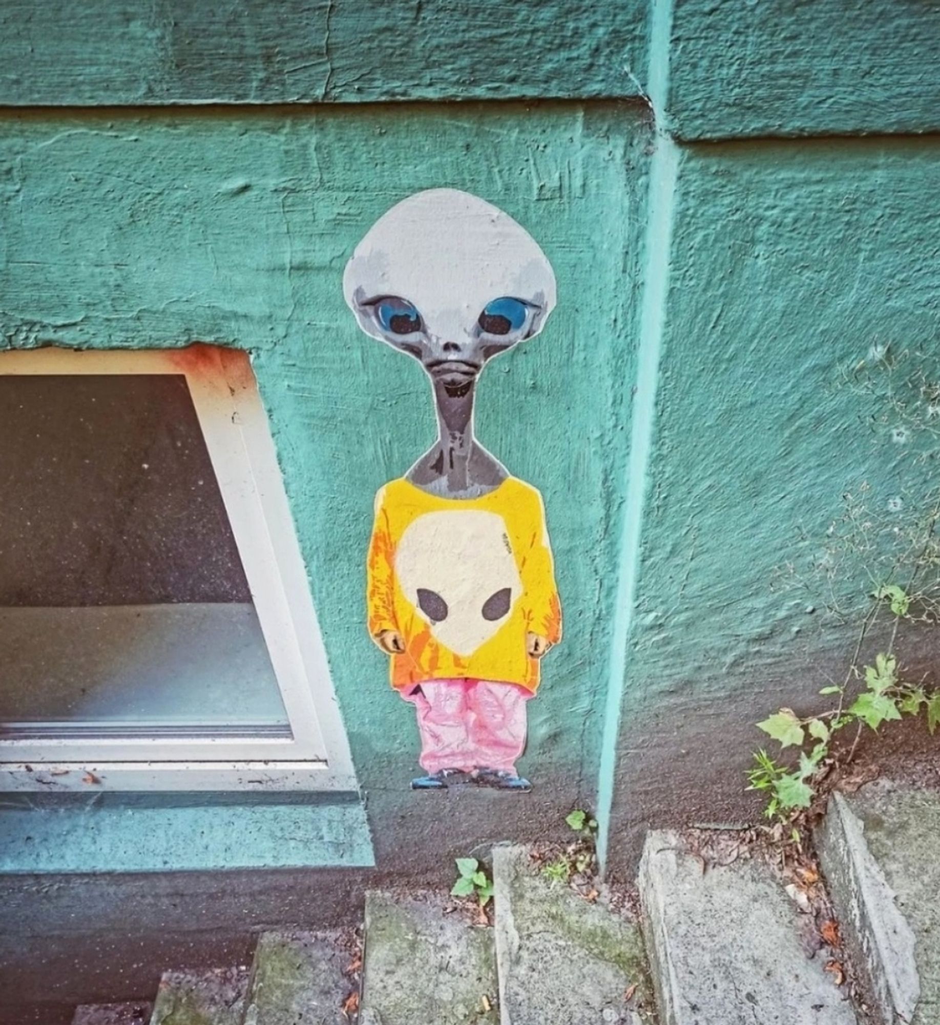 Streetartwall. On a green-painted wall next to a white basement window and above a basement staircase, there is a paste-up with a fashionable alien. The small paste-up is colorful because the gray alien with blue eyes is wearing a yellow sweater with an alien head on it. It has chosen a daring combination with pink baggy trousers and blue sneakers. Judging by the somewhat sad expression on its face, there is still room for improvement.