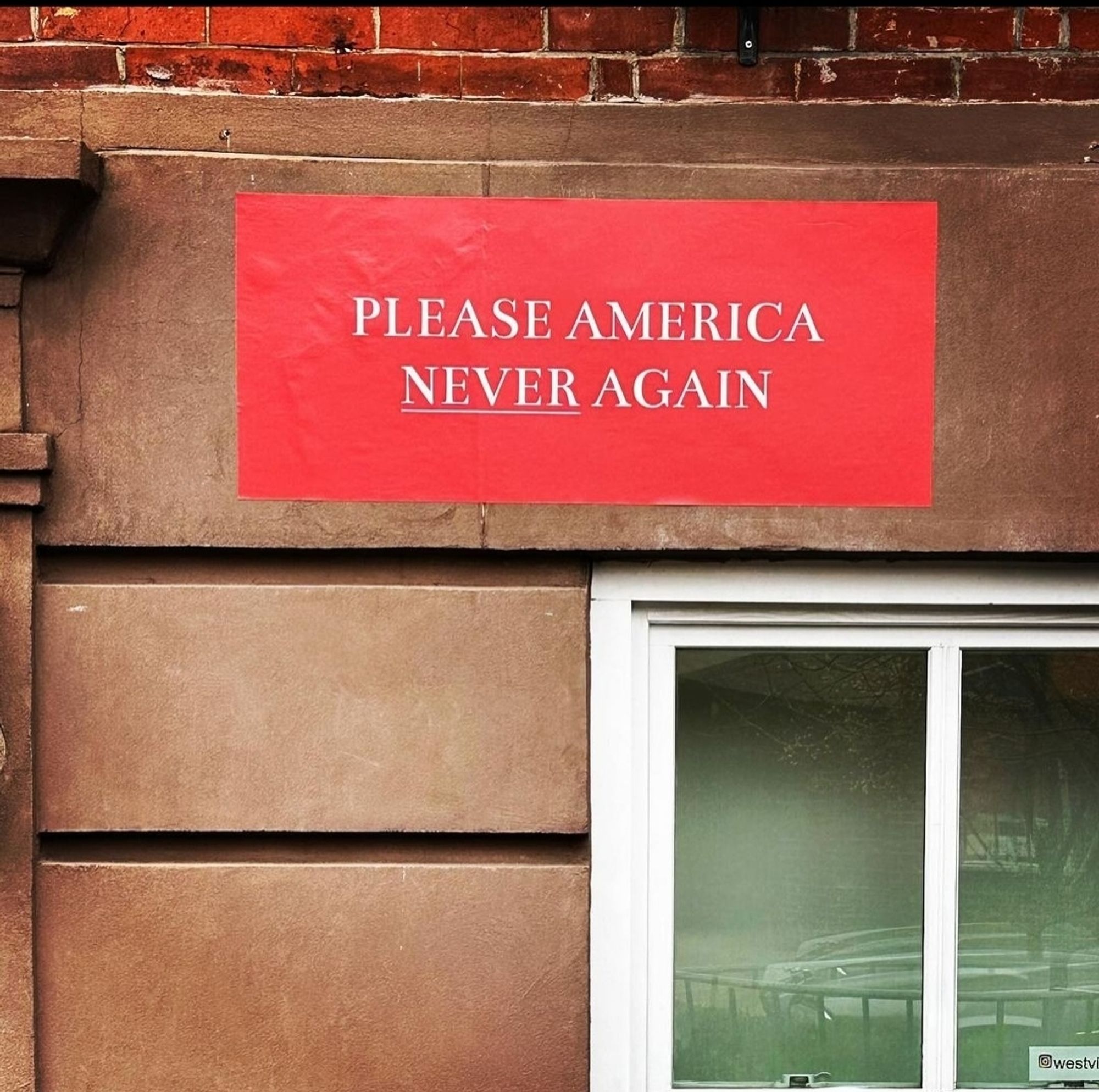 Streetartwall. A red poster with white text is attached to a brown wall with a window. It reads in capital letters: "Please America never again". The word "never" is underlined. 
Info: The sentence refers to the presidential elections in the USA.
WestvillageFamous is an anonymous designer who sticks signs with messages on the wall of his house in New York and then posts them on the Internet.