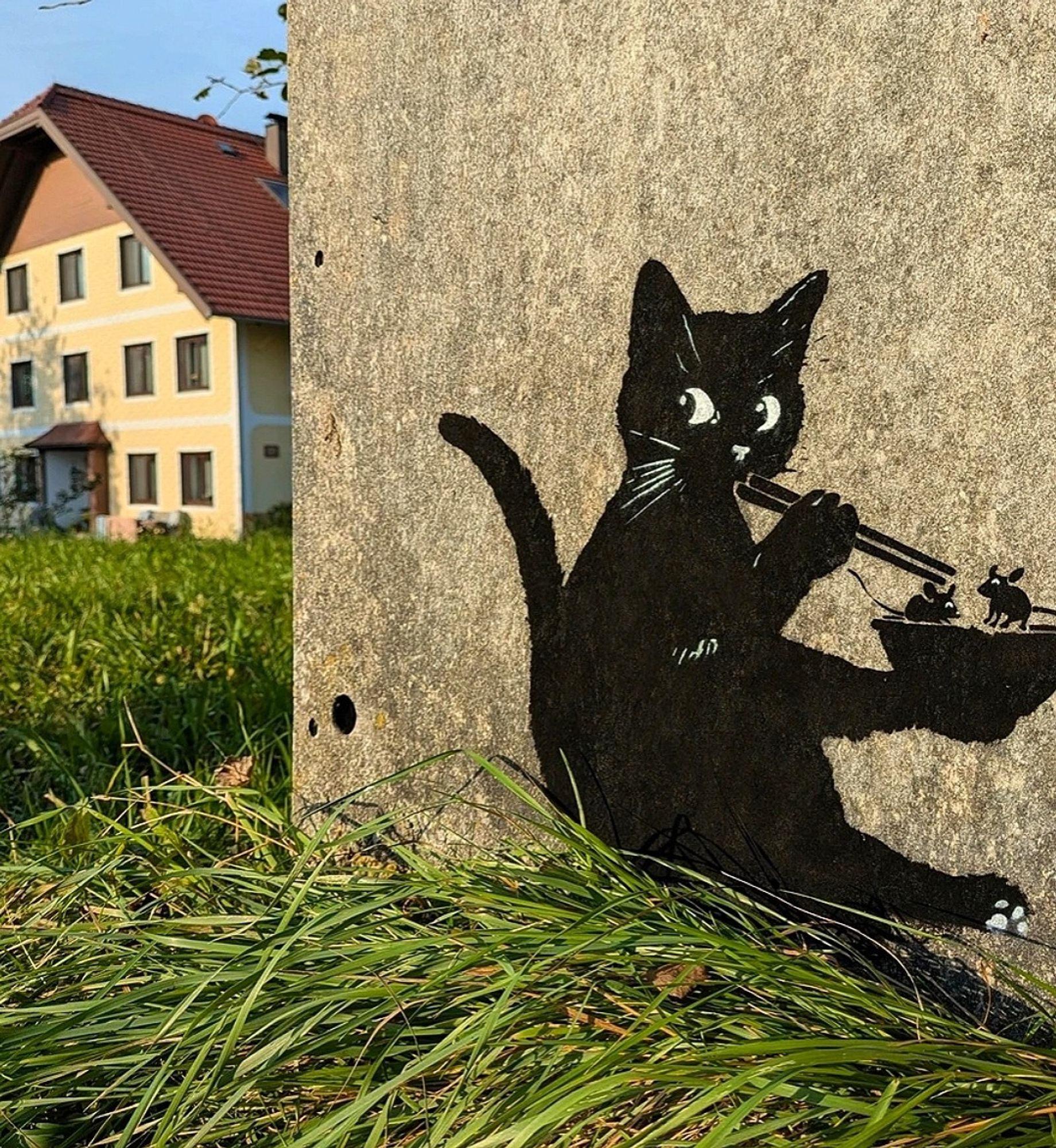 Streetartwall. A small stencil mural with a black cat was sprayed/painted on an old beige-colored house wall near the ground. The cat sits there with a bowl in one paw and holds the sticks with the other paw. It looks around furtively. Two black mice are sitting in the bowl and look very frightened.