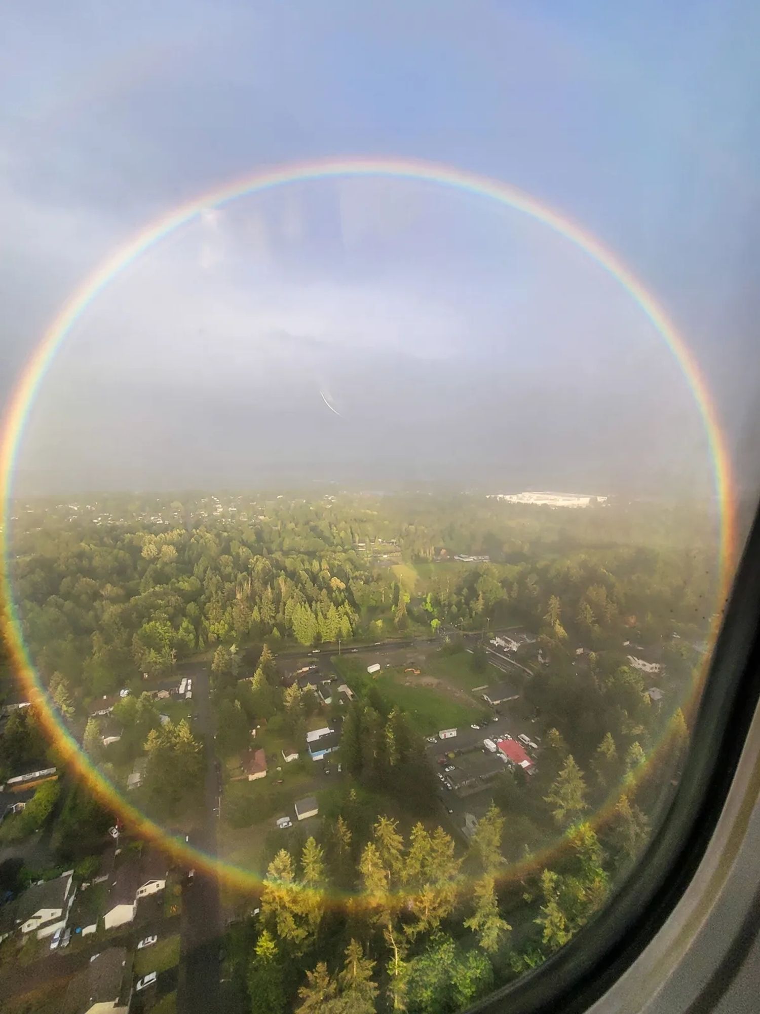 Photography.  A color photo of a circular rainbow, taken from an airplane window on landing. The view is over an urban landscape with many trees and a slightly cloudy sky. A shimmering rainbow shines over everything, but not as a semicircle on the horizon, but as a full pastel-colored circle over the entire scenery. A rare sight, made possible by the right angle (between 42° and 41°), the weather conditions, the perspective, the height and, of course, a lot of luck.
Info: 
Photographer Peter Reinold held his camera phone steady through a turbulent landing at Seattle-Tacoma International Airport to capture this complete circular rainbow: "I always pick the window seat on planes, as you never know what you [might] encounter. It paid off this time."