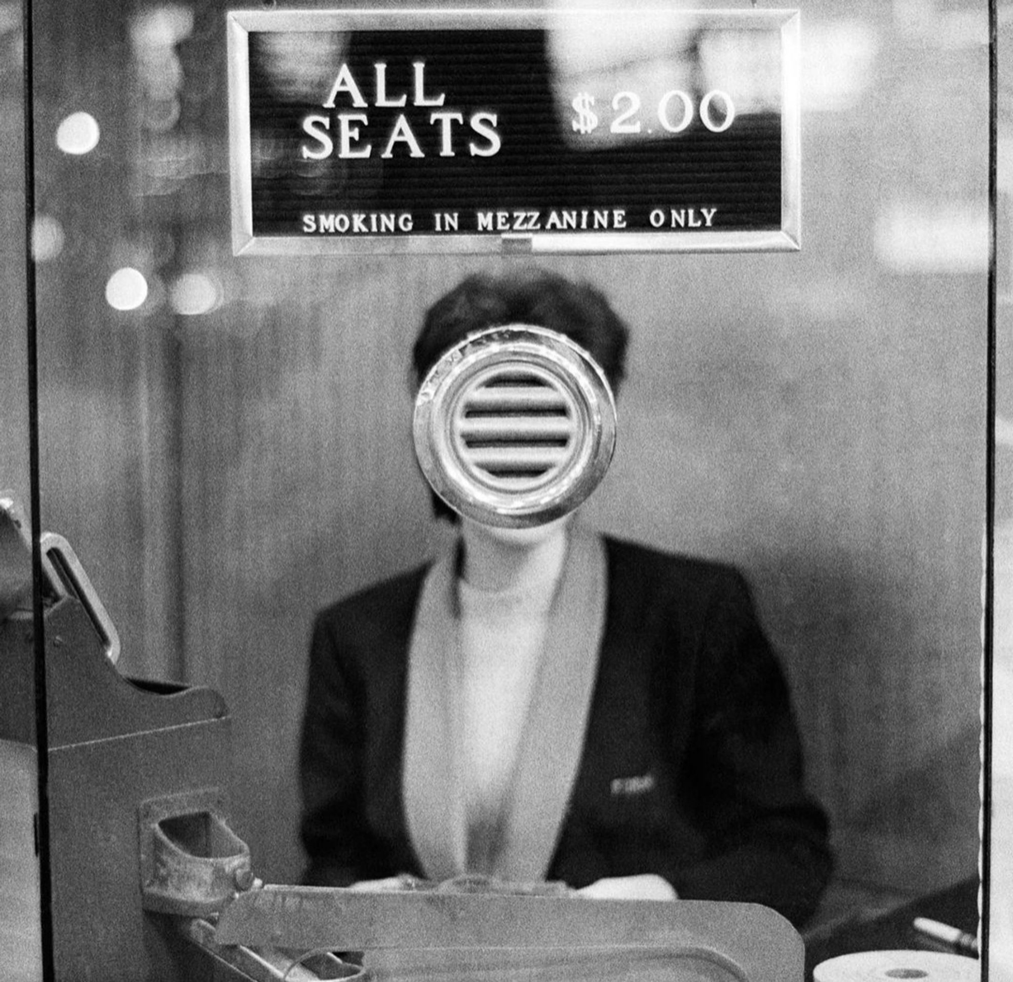 Photography. A black and white photo of a cashier in the entrance area of a theater. The cashier's face, standing in the center of the image, is completely obscured by the small metal intercom window through which one would speak to her, giving the lady in the black costume a surreal faceless appearance. Above her hangs a sign that reads, "All seats $2.00 - Smoking mezzanine only" 
Info: Joel Meyerowitz (born 1938) is one of the most important contemporary photographers in the world. He began with black and white street photography in the Bronx in the 1960s and was a pioneer and important trailblazer of color photography in the 1970s.