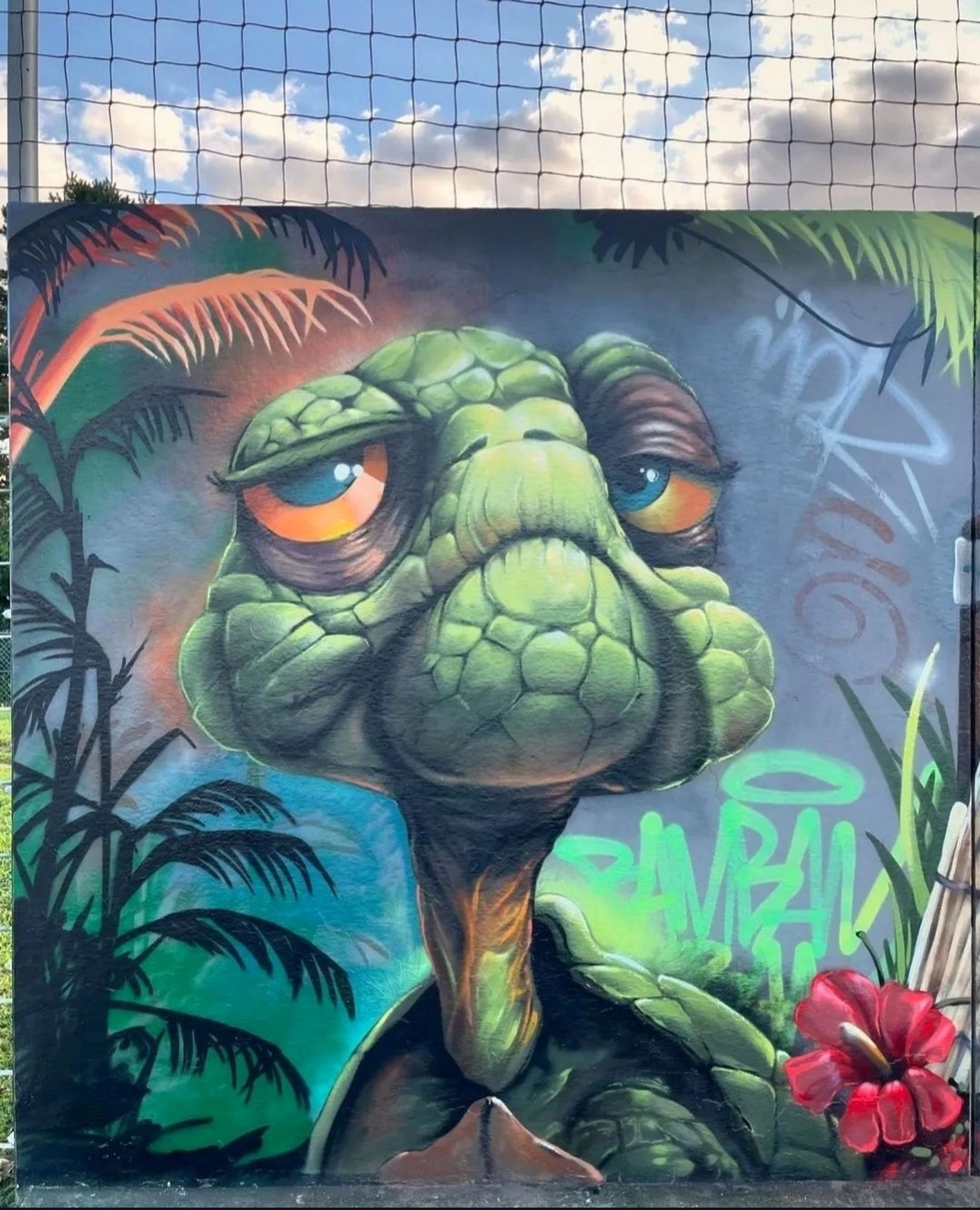 Streetartwall. The head of a friendly turtle was sprayed/painted in comic style on a long street wall with a wire fence. The cute animal is surrounded by a jungle of green ferns, leaves and a red orchid. She has short shoulders, a thin long neck and big orange googly eyes. Her bedroom eyes will make you melt away.