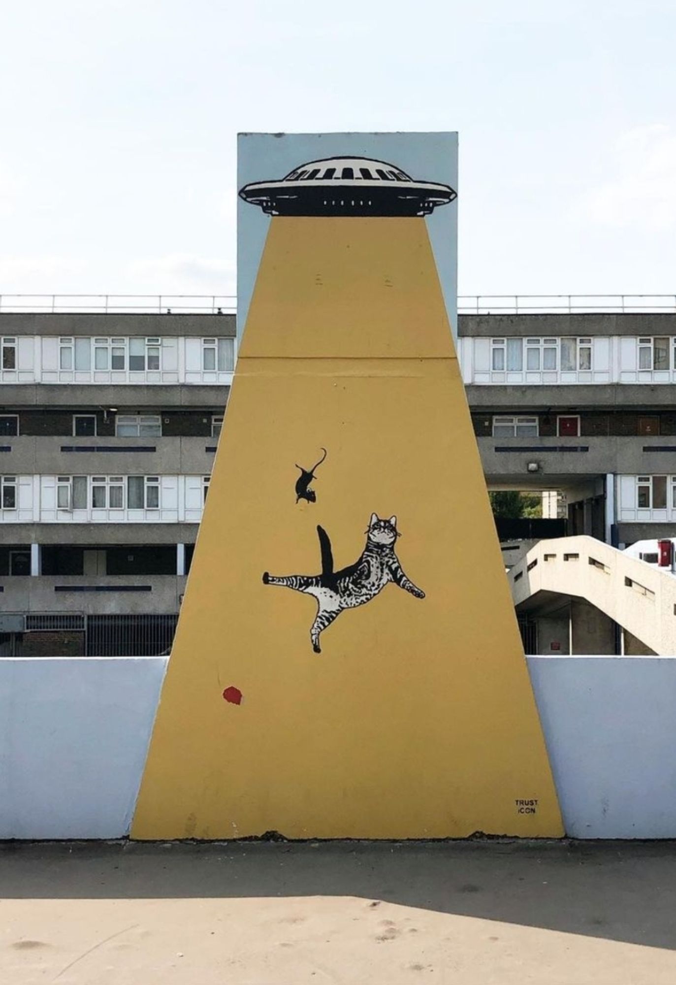 Streetartwall. A funny comic mural of an alien invasion has been sprayed onto a narrow three-storey wall that widens downwards to form a triangle. A UFO can be seen in the upper part of the tower, the rest of the triangular wall is the yellow transporter beam. Trapped in this yellow light are a striped cat and a rat floating upwards through the air.