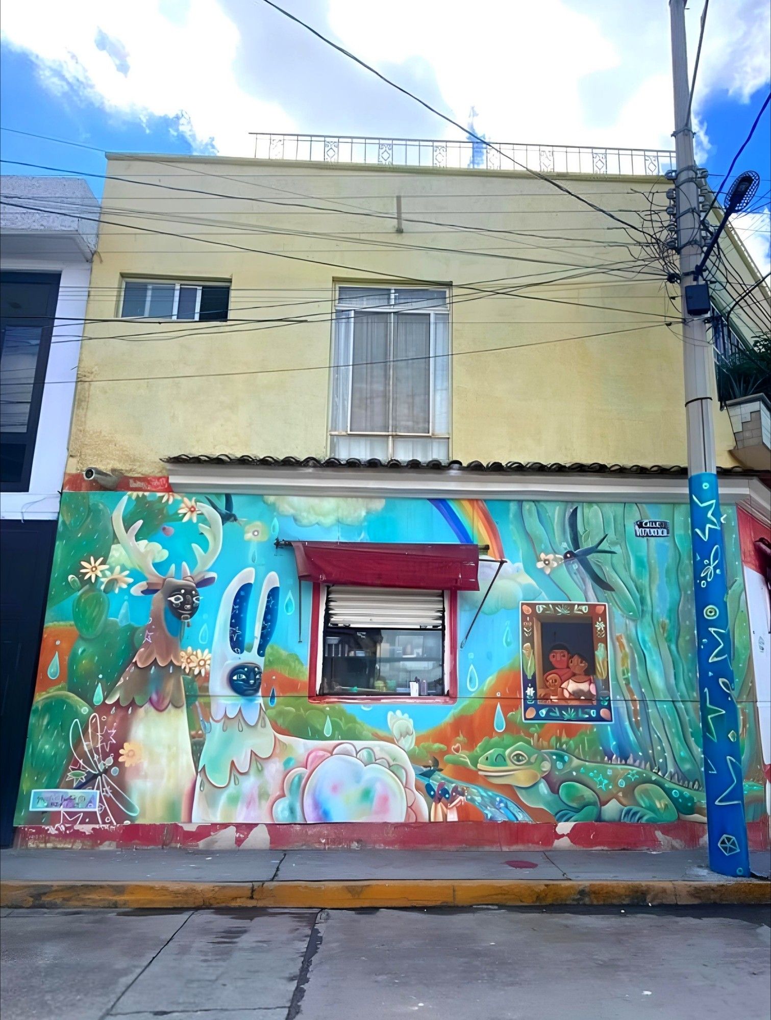 Streetartwall. On the façade of an old Housewares store in Mexico is a beautiful naive mural with a family and colorful fantasy figures. The whole wall is painted in bright colors and shows two large, fluffy animals on the left side. They look like alpacas, but one in brown with antlers and one in pale rainbow colors with bunny ears. Next to them sits a kind of green frog lizard, birds fly with flowers through the green landscape. A rainbow can also be seen. A family with a father, mother and child can be seen in a small ornate brown window. A pretty mural in typical naïve folk art, but painted here by a Japanese woman at a street art festival.