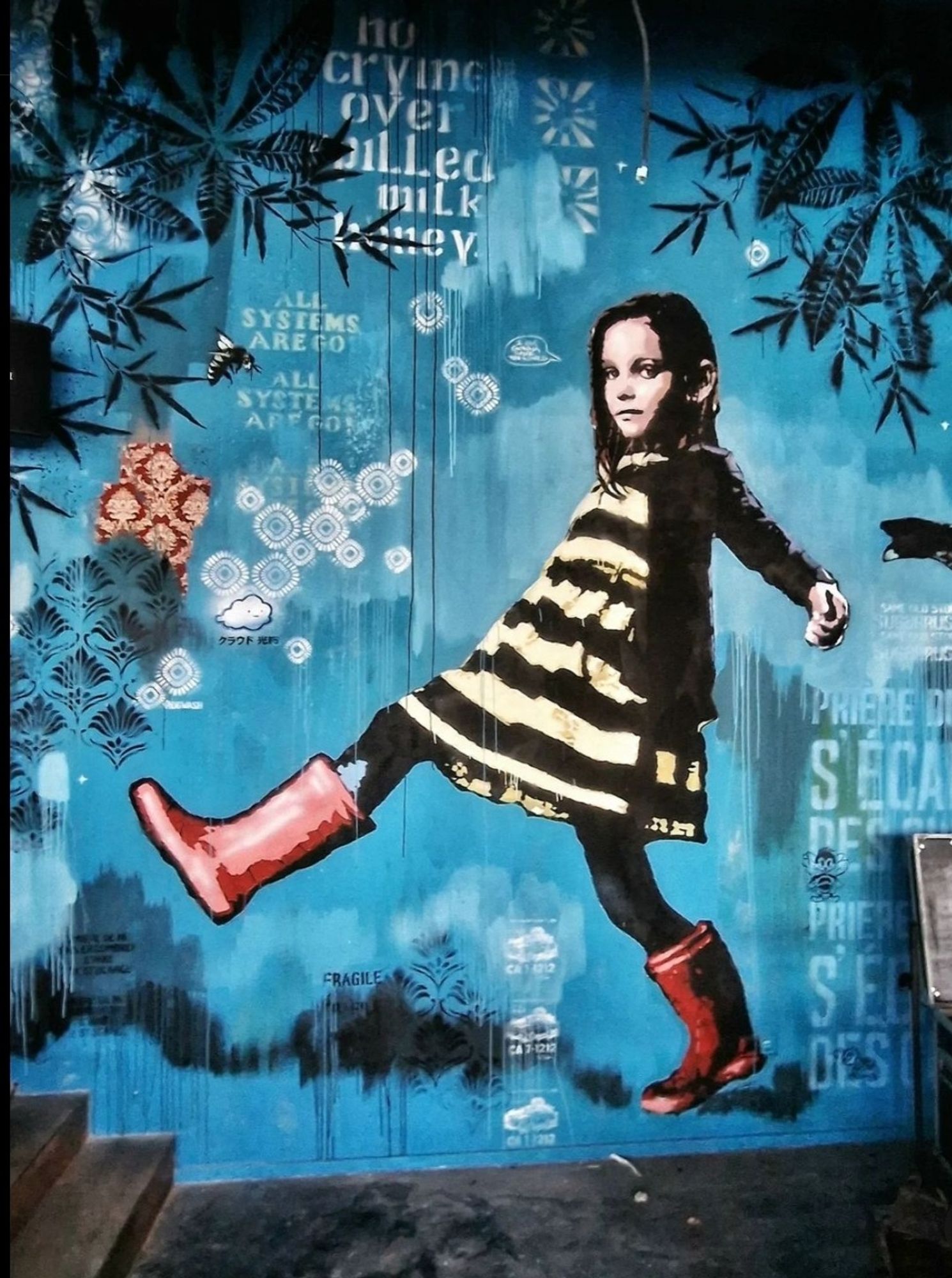 Streetartwall. The mural of a running girl was sprayed, glued and painted on a blue painted wall next to a black staircase. The background is decorated with a collage of small star-shaped patterns like pieces of wallpaper and small texts. There are leaves and bees at the edges. In the center, a dark-haired girl in a black and white striped dress, black tights and red rubber boots walks across the wall. One leg is in the air, her arms back as if she is balancing through a puddle. She is looking at the viewer. Note: The small texts are difficult to make out in the photo. Some of them are: "No tears over filled milk honey" / "All systems are go" / "Ca 7-1212".
Info: Pipsqueak was here!!! is an Amsterdam based artist duo, driven by a strong passion for all life on planet earth.