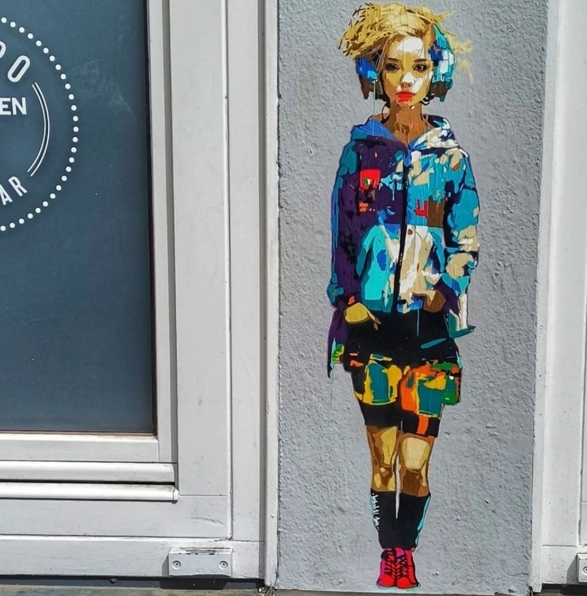 Street art wall. On a gray wall between two windows is a small comic-style mural depicting a young woman. The small mural is wonderfully colorful and shows a standing young woman with a short mop of blonde hair, blue headphones and dressed in a colorful rain jacket, matching shorts, black stockings and red boots.